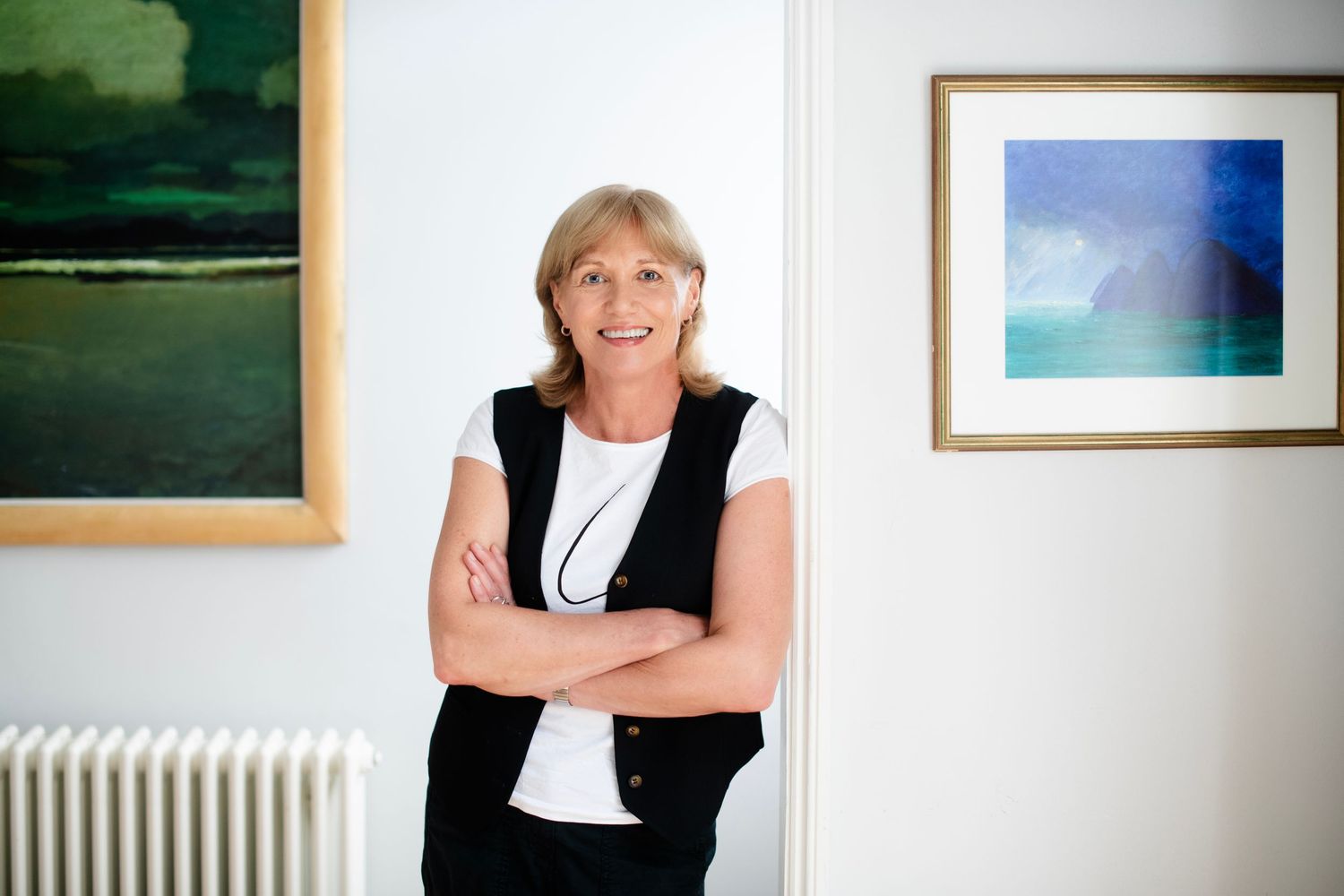 Smiling woman with short blonde hair stands with crossed arms near artwork in a bright white gallery space.