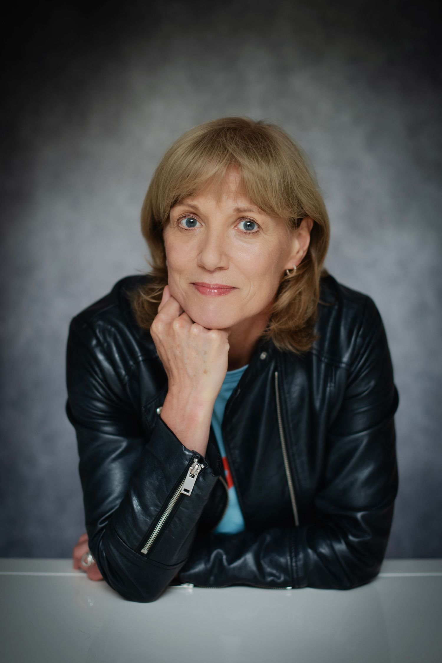 Woman in black leather jacket resting chin on hand, gazing at camera against dark gray background.