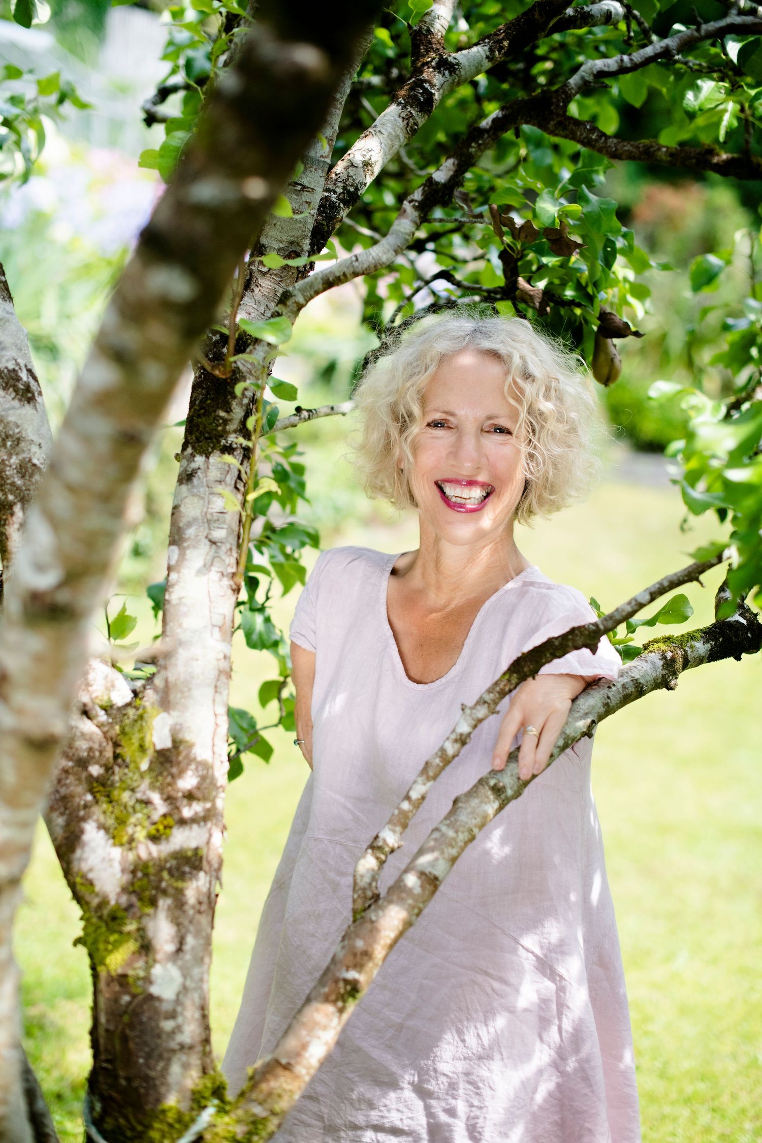 A laughing woman with short curly blonde hair stands among green tree branches in a sunny garden wearing a pink dress.