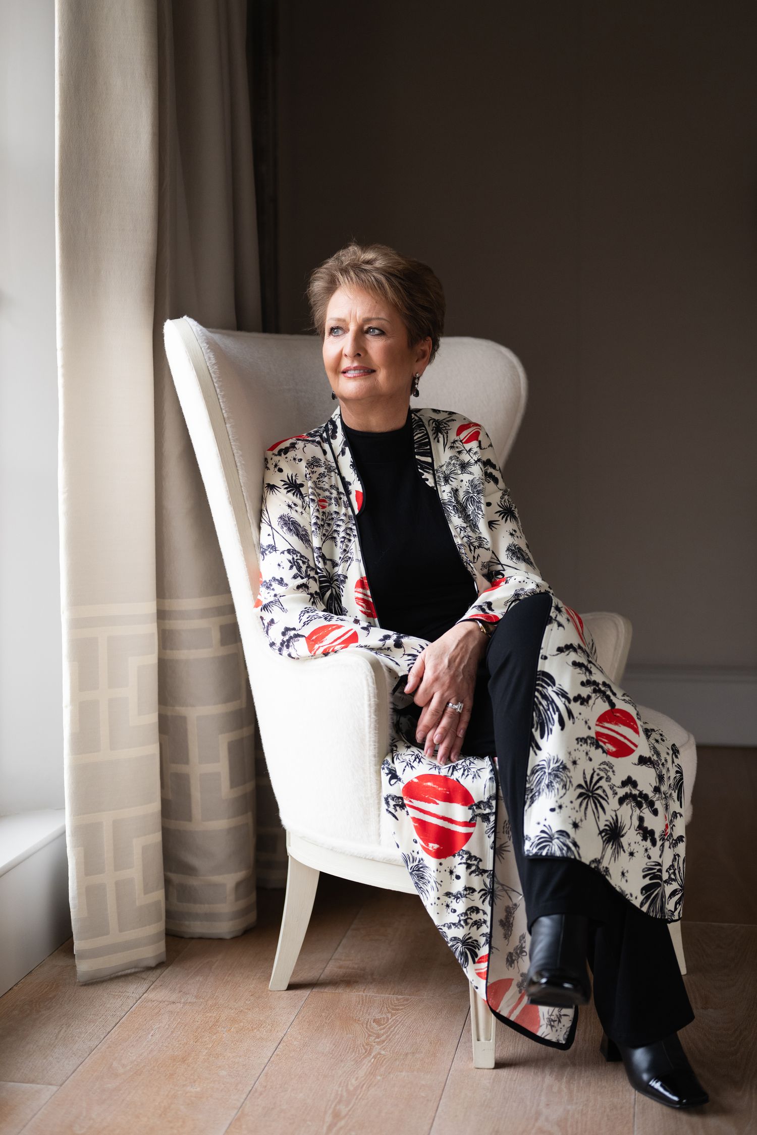 Elegant woman in floral coat seated in white armchair near a window in stylish room.