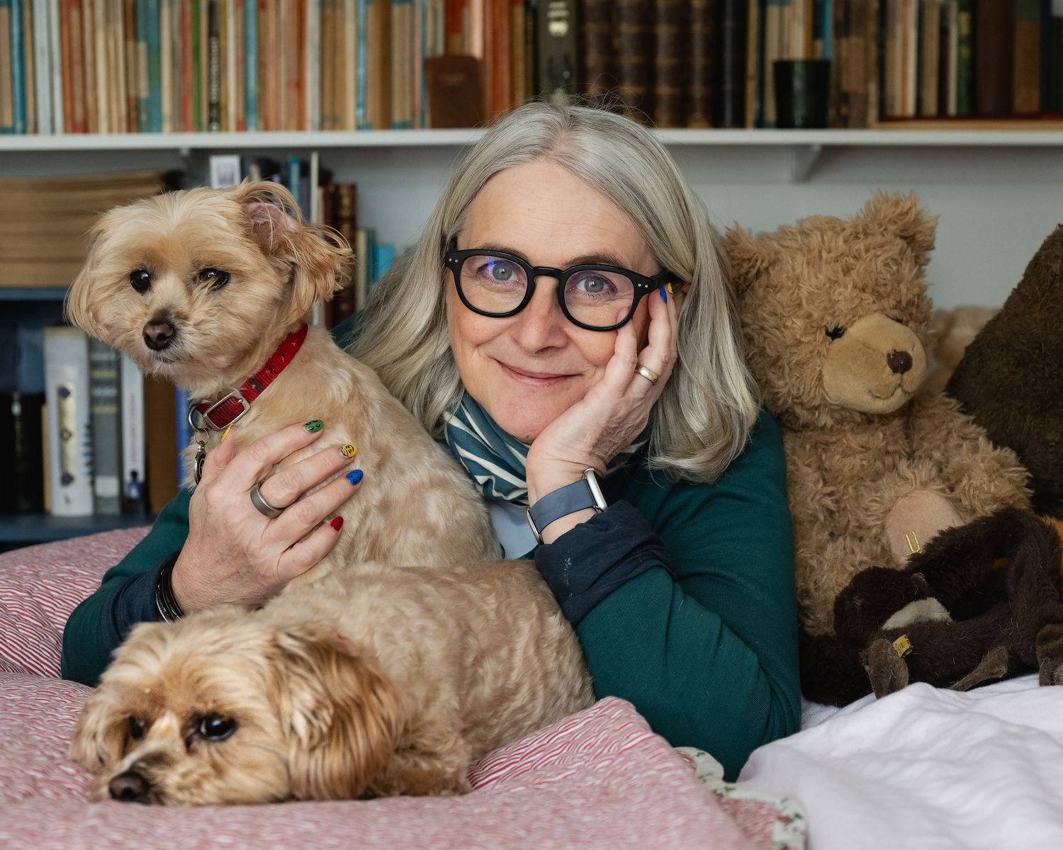 Smiling woman with white hair lying with two small dogs and teddy bear.
