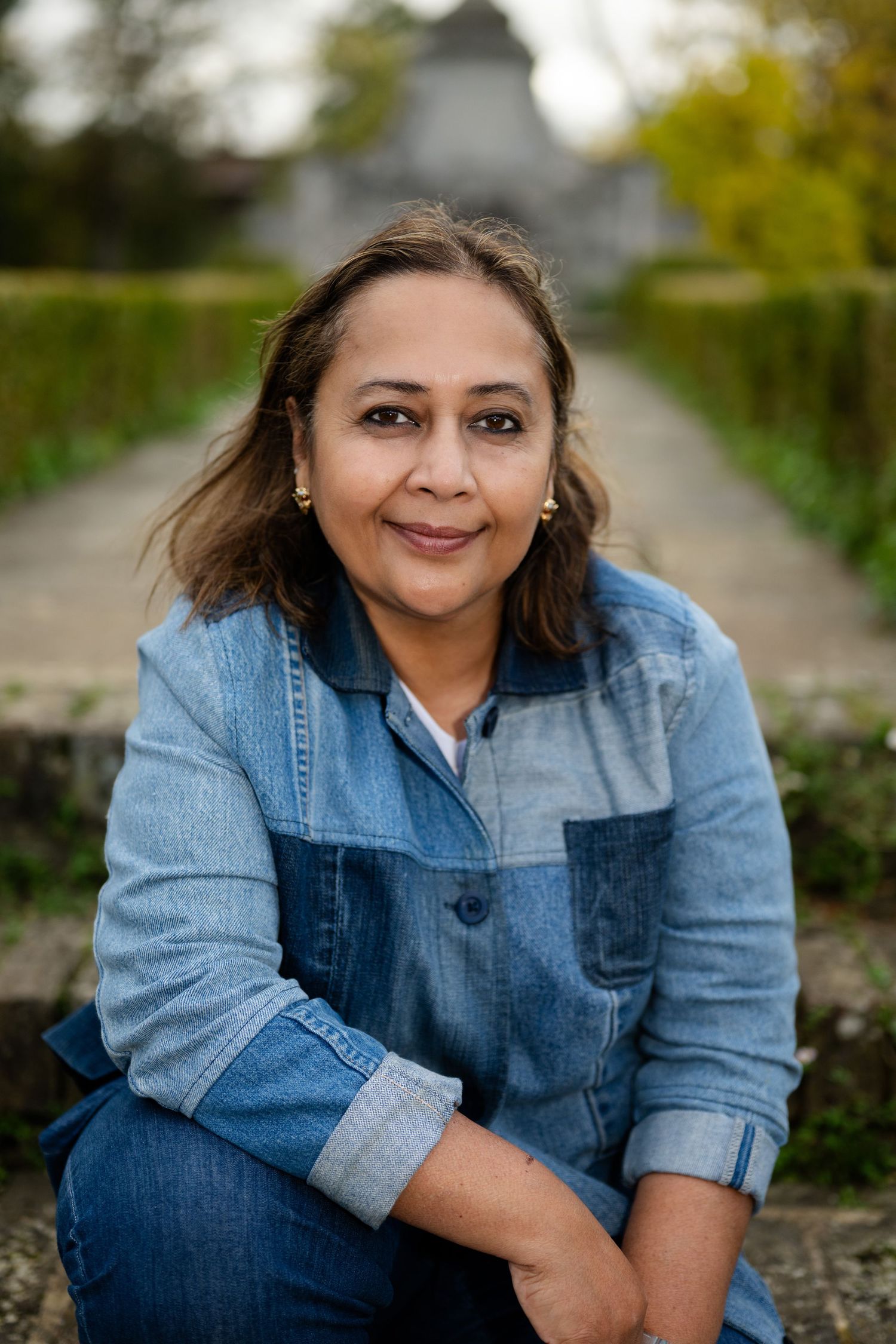 Close-up portrait of a smiling woman in denim shirt sitting outdoors in a garden setting.