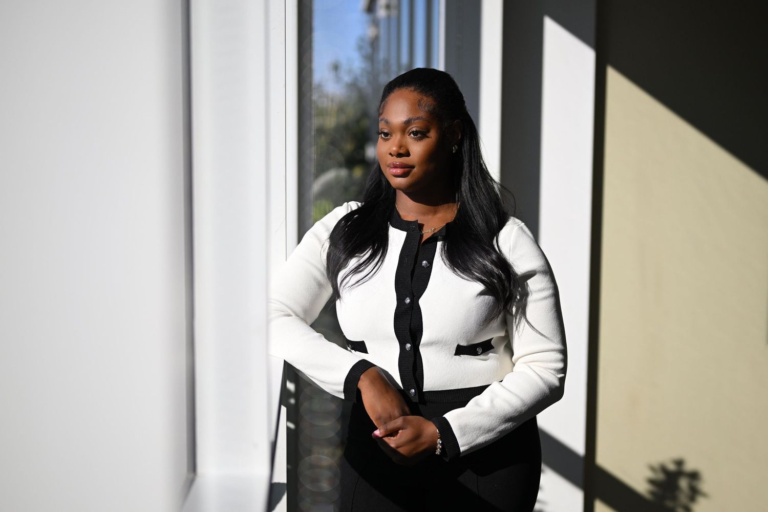 Professional woman in white ribbed jacket standing near sunlit window indoors.