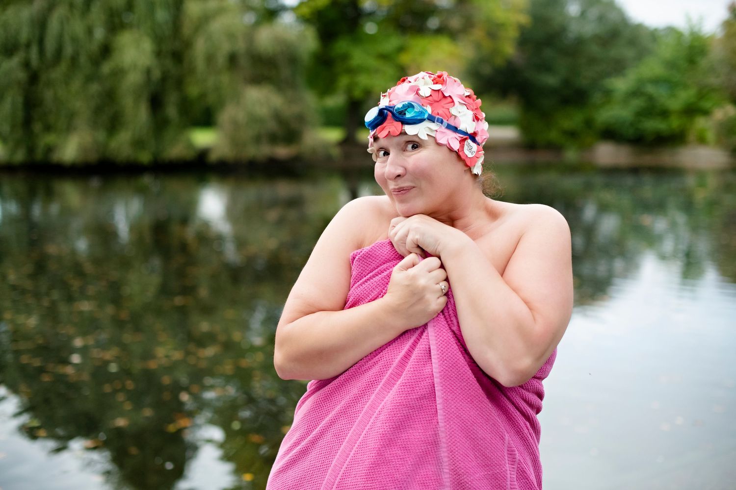 Woman in pink towel and floral swim cap with goggles standing by a lake after swimming outdoors.