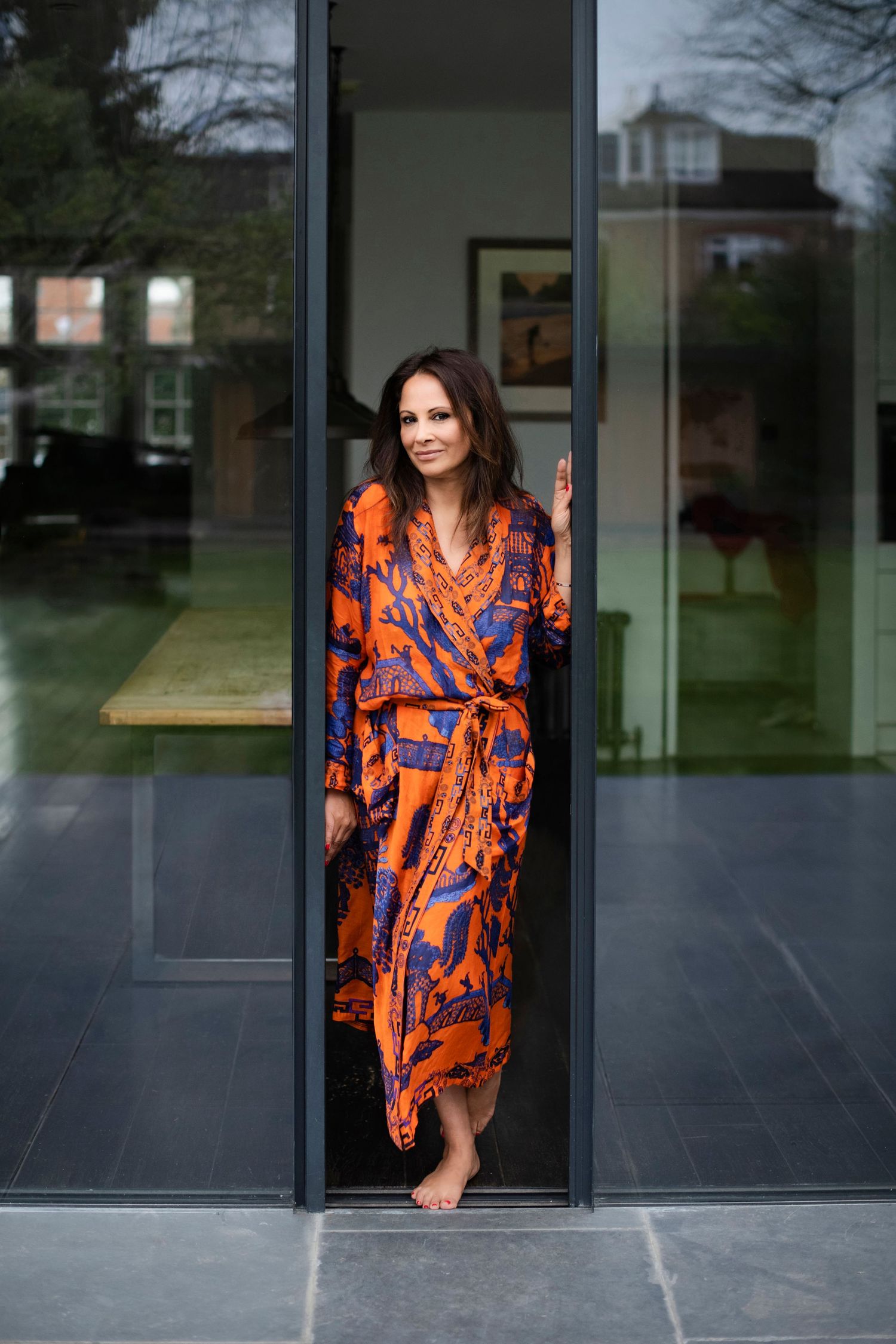 Woman in vibrant orange and blue patterned robe standing barefoot in modern glass doorway.