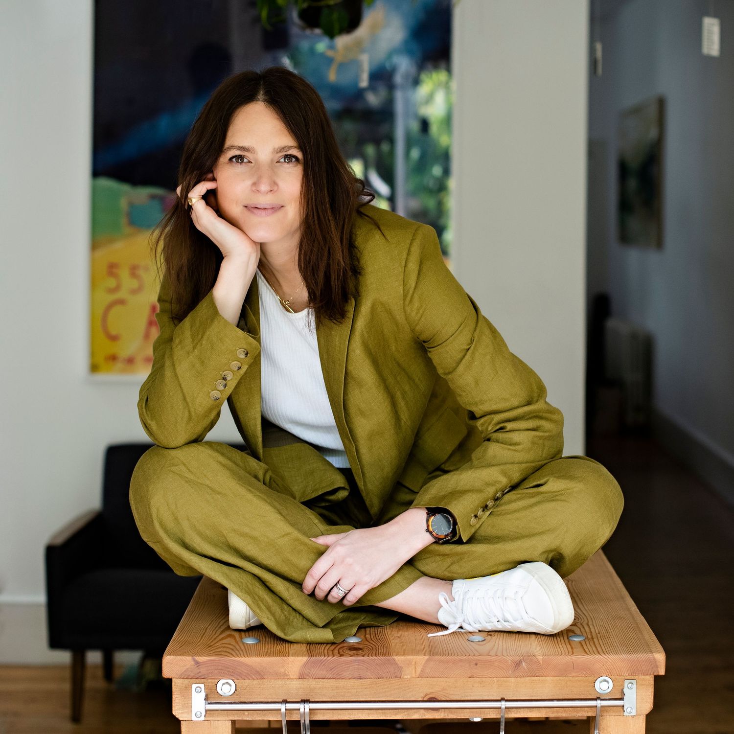 Woman in olive suit sits cross-legged on wooden table in bright modern interior.
