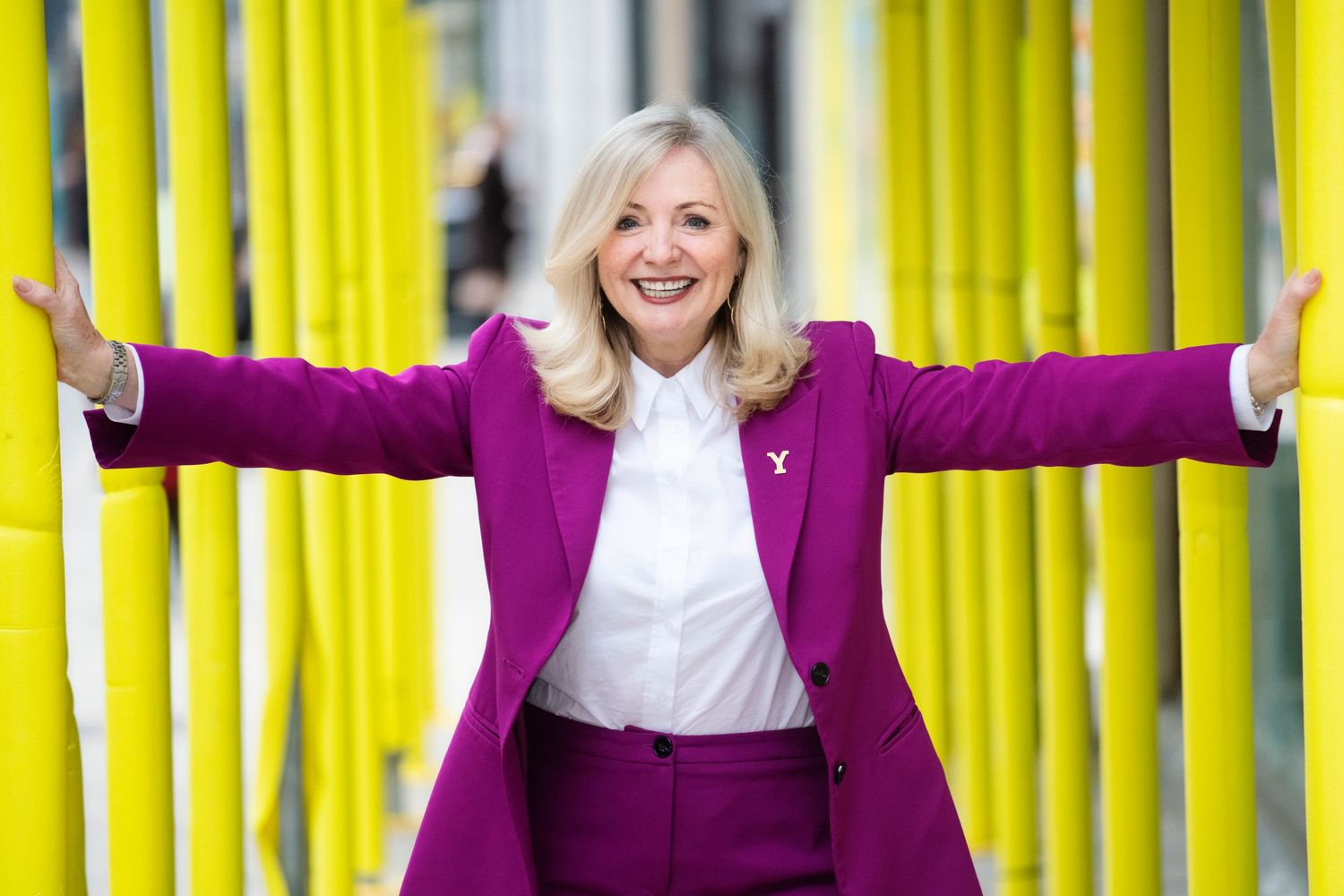Enthusiastic woman in purple blazer stands with arms wide open near yellow bamboo.