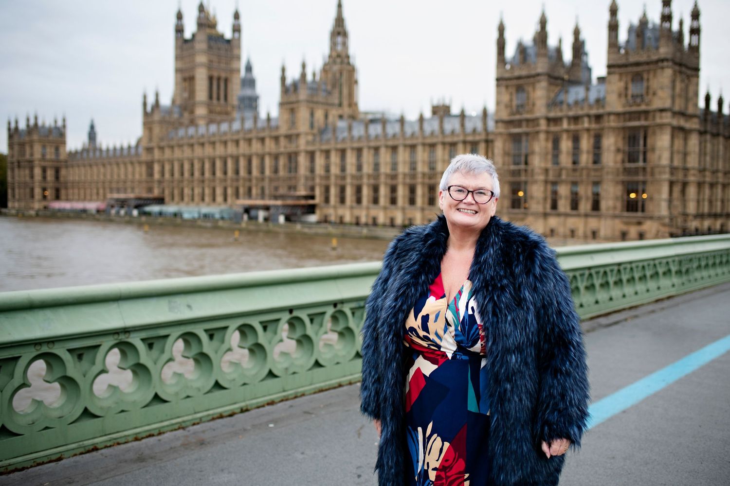 Smiling woman with gray hair and glasses in a black fur coat standing on Westminster Bridge in London.