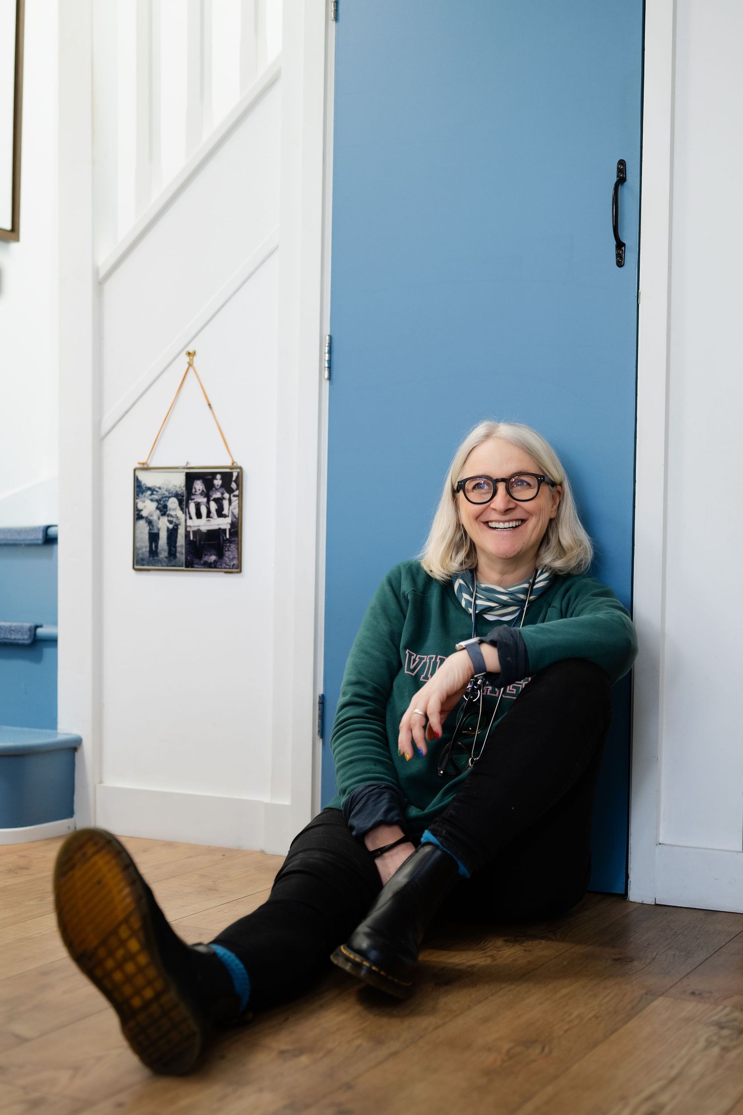 Cheerful woman with gray hair and glasses sits on wooden floor leaning against a blue door.