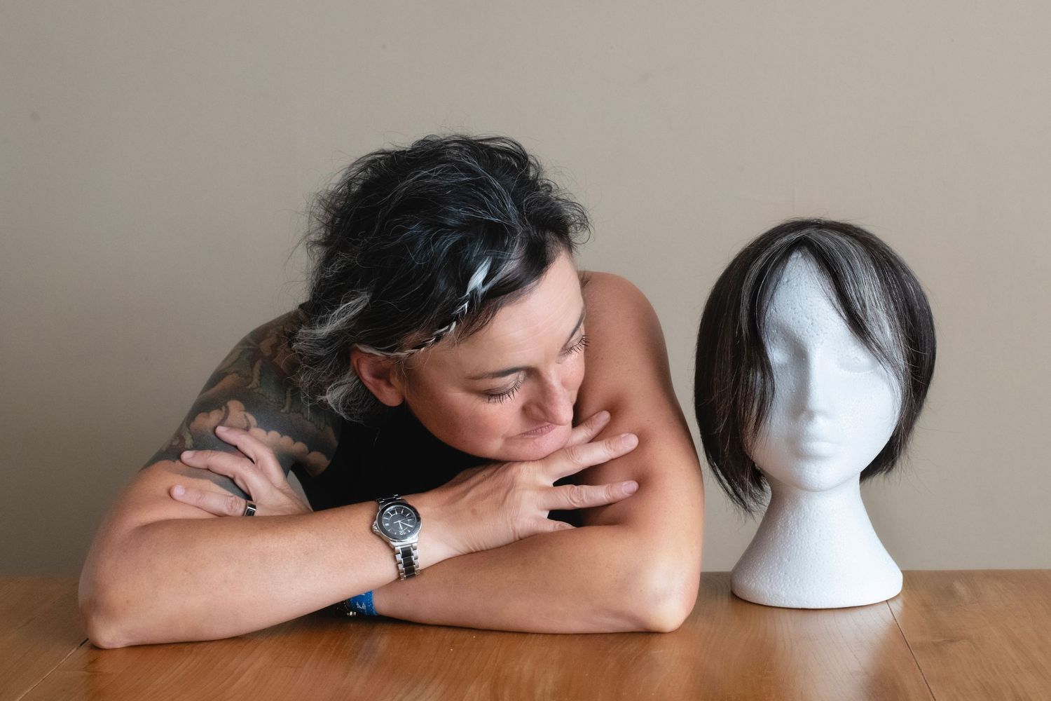 Woman with tattoos leaning on table beside wig on mannequin head.