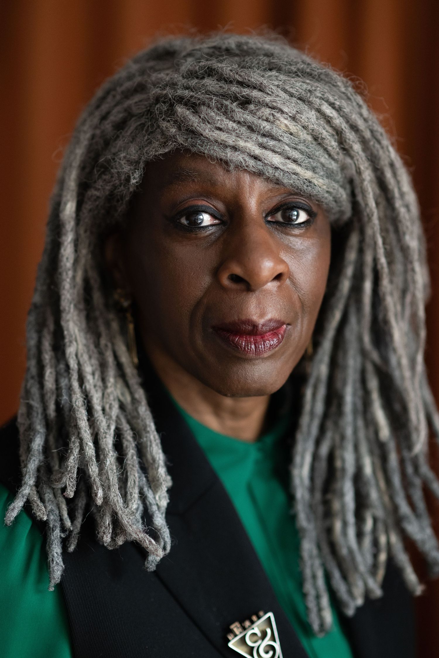 A striking Black woman with long gray dreadlocks wearing a green top and black blazer gazes intensely.