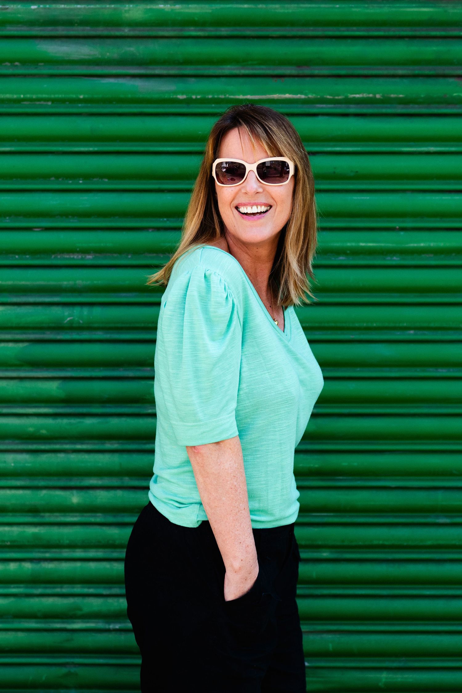 Smiling woman in mint top and sunglasses posing against green corrugated metal wall.