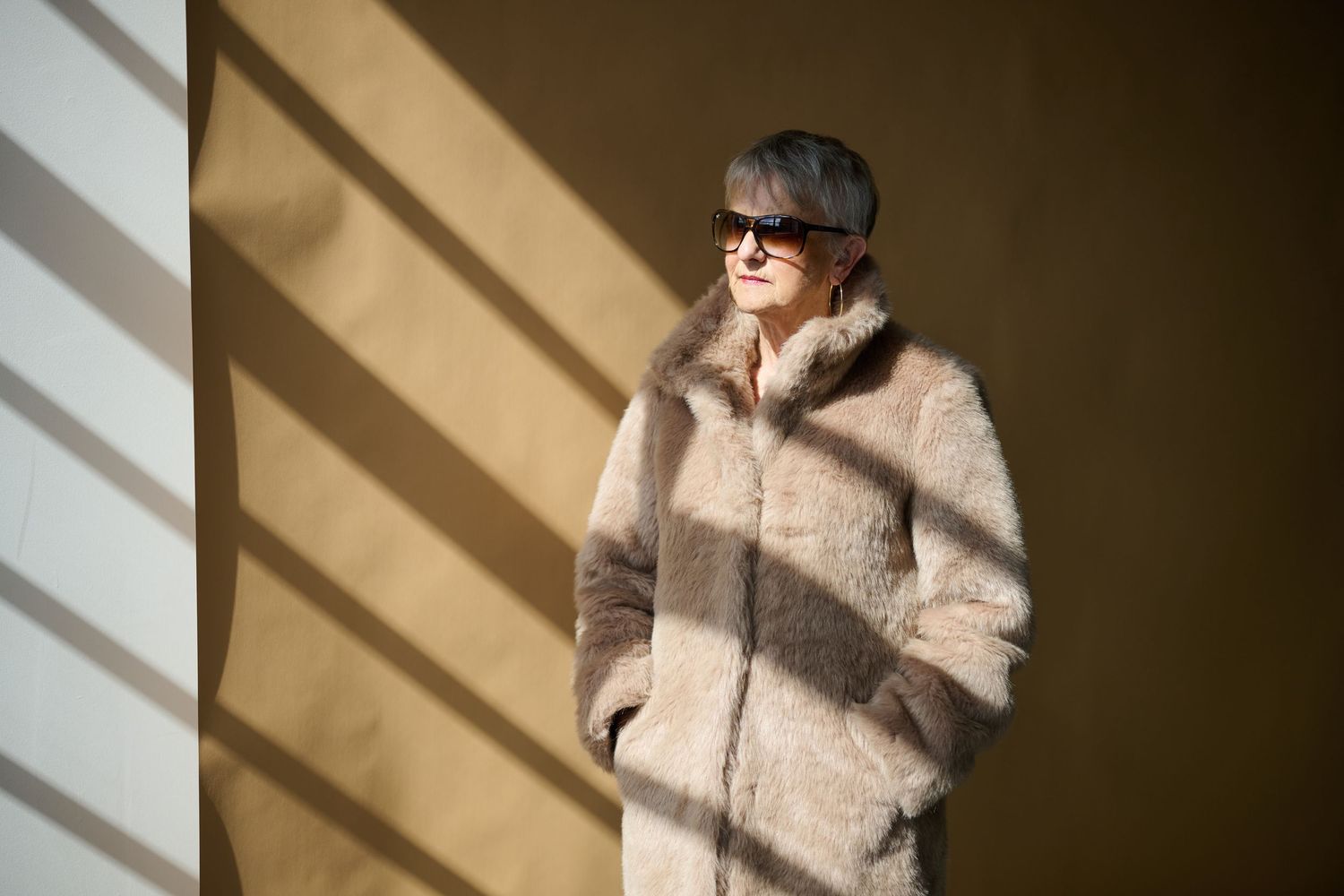 An older woman in a beige fur coat and sunglasses stands in warm sunlight with shadow patterns.