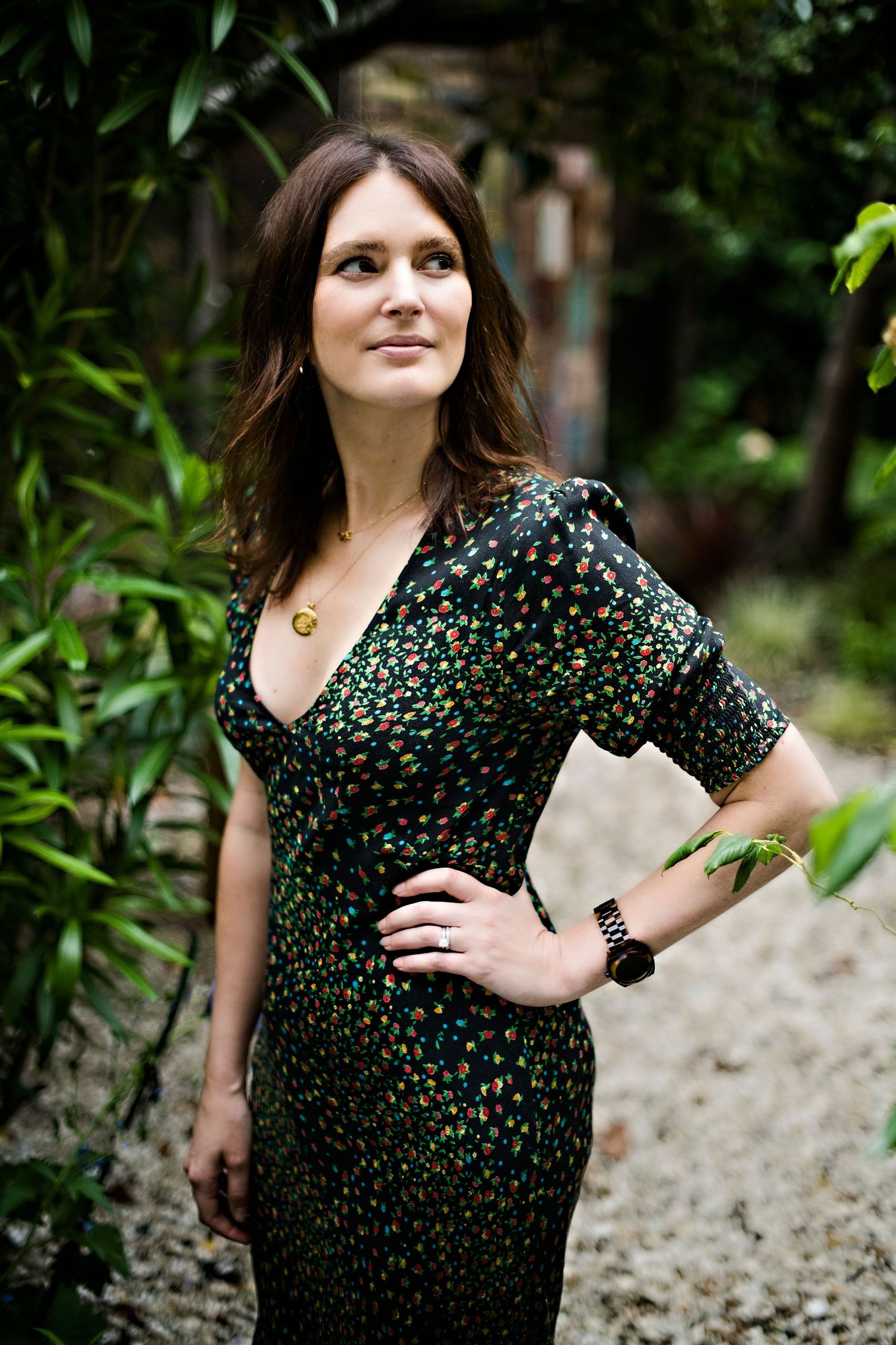 A brunette woman in a floral dress poses confidently in a lush garden setting with bamboo plants.