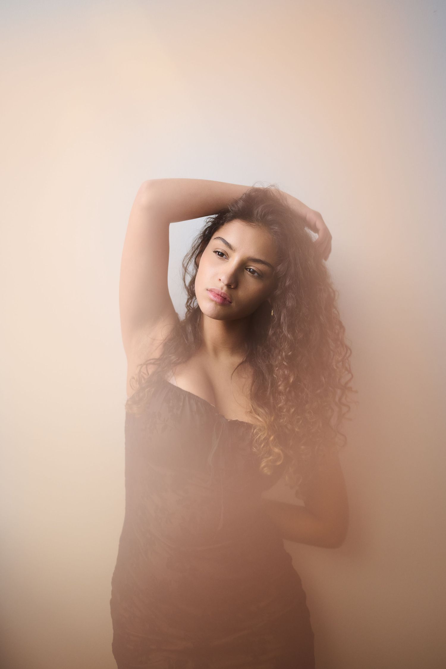 Young woman with curly hair in a black dress posing with arm raised in soft foggy light.