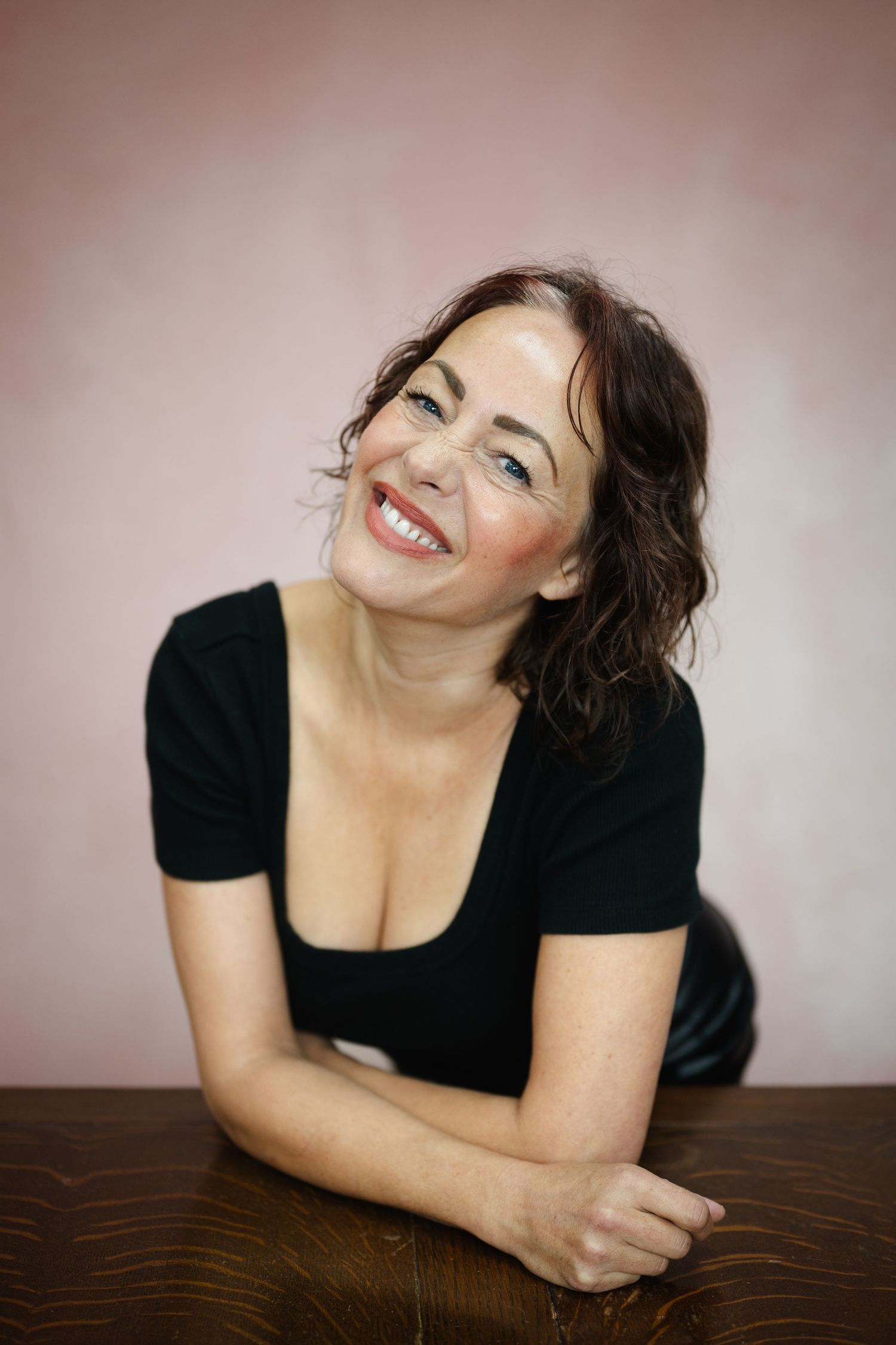 Smiling brunette woman in black top leaning on wooden table against a soft pink background.
