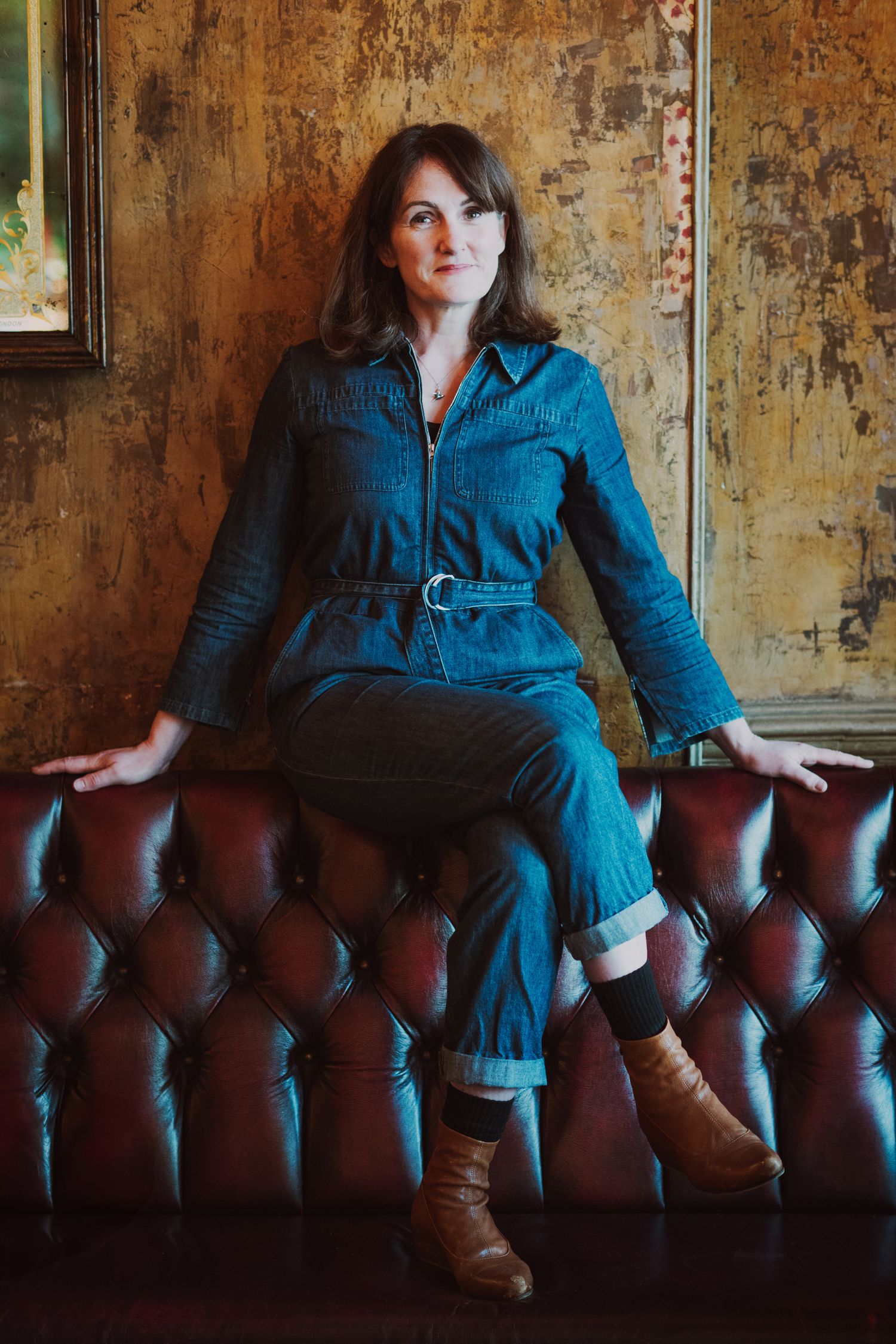Woman in belted denim jumpsuit seated on tufted leather sofa against rustic distressed wall.