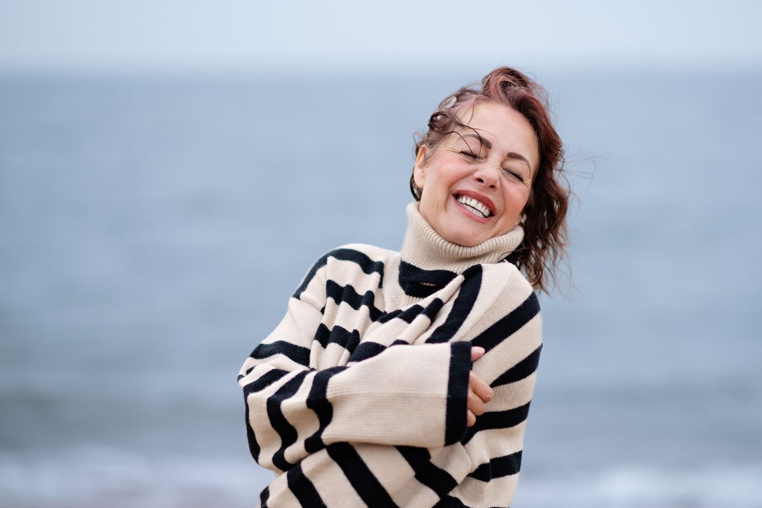 A cheerful woman in a striped turtleneck sweater hugs herself while laughing on a beach.