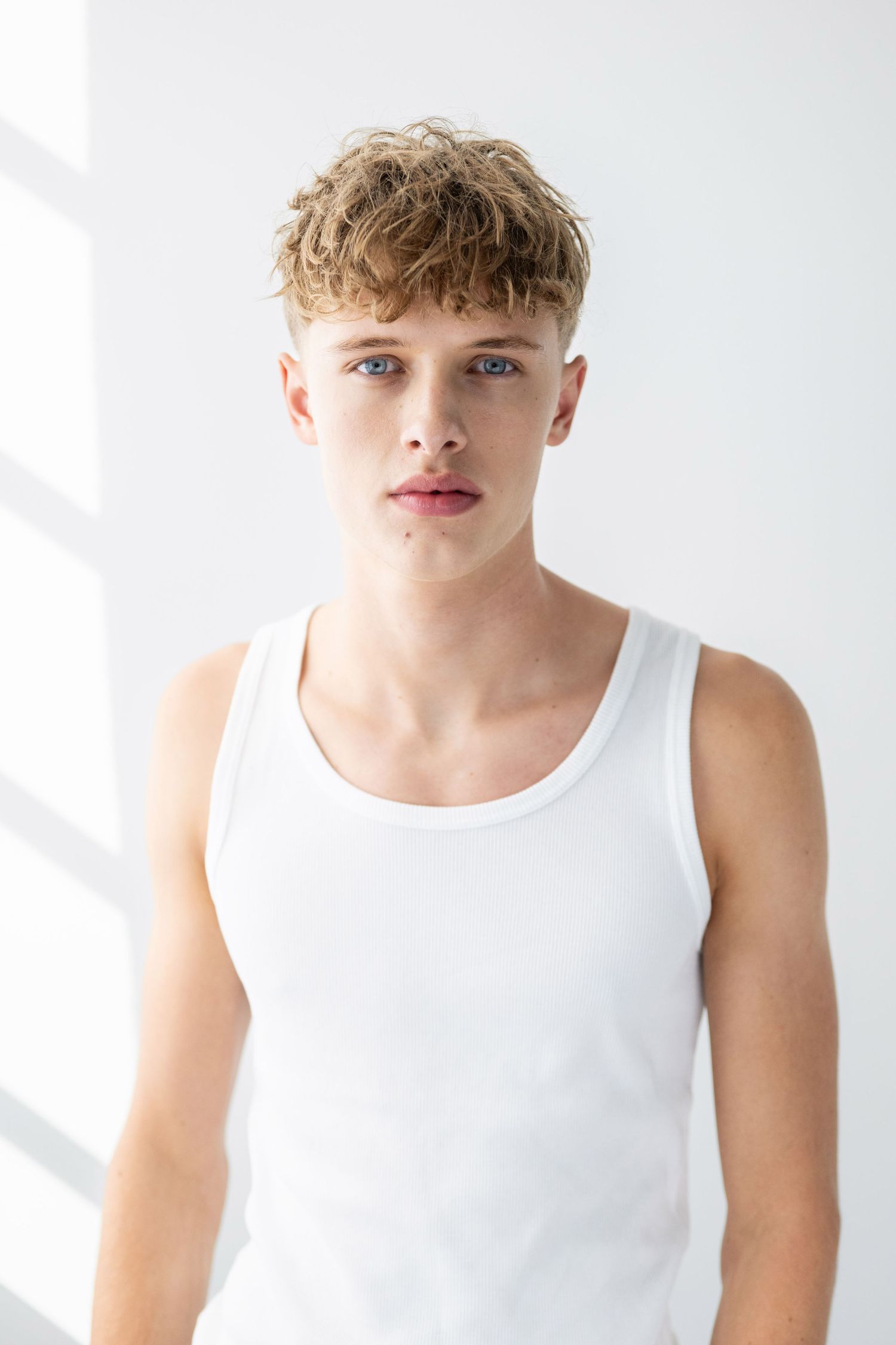 Young man with curly blonde hair and blue eyes wearing a white tank top against a white background.