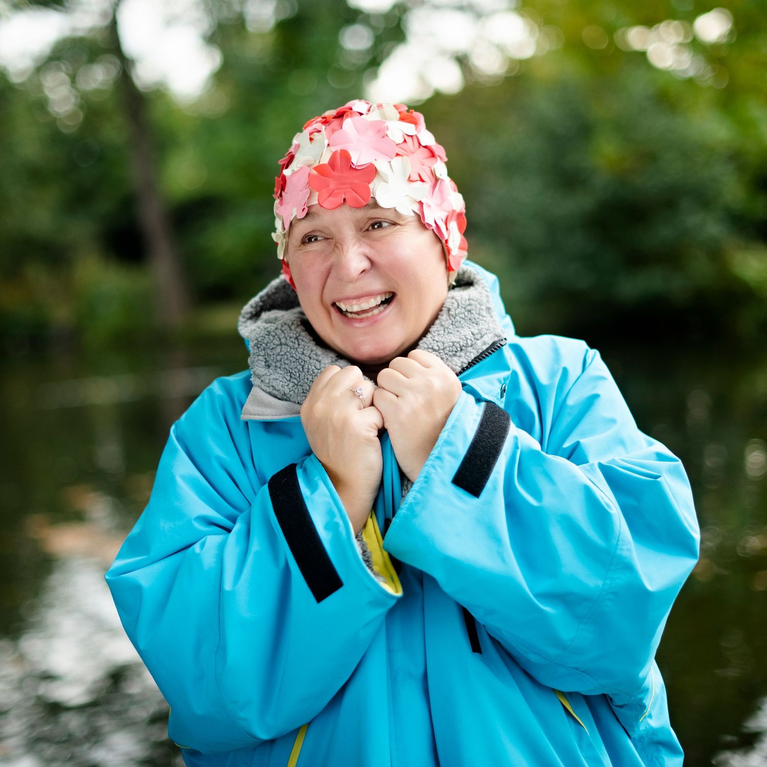 Smiling woman in blue jacket and floral swim cap standing near a lake outdoors.