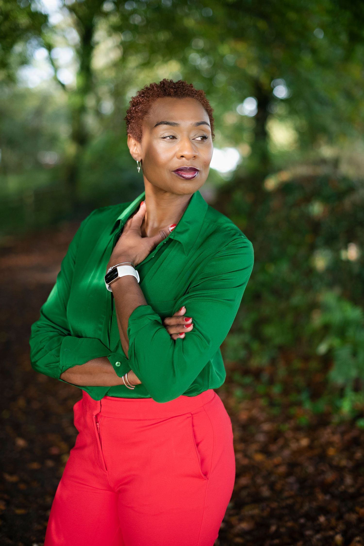 Black woman in green shirt and red pants posing thoughtfully with arms crossed in a lush green outdoor setting.