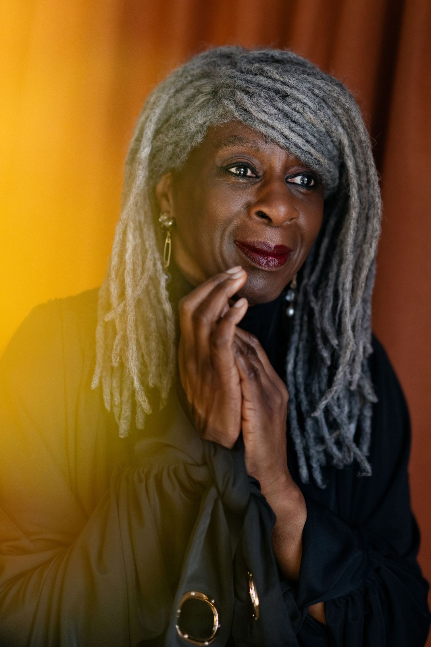 An elegant older Black woman with long gray locs poses thoughtfully against a warm golden background.