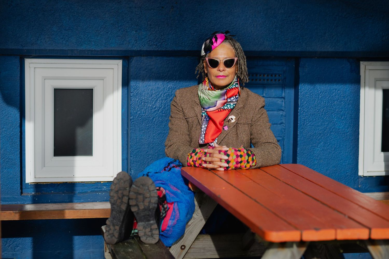 A fashionable woman with colorful braids and sunglasses sits at a wooden picnic table by a blue wall.