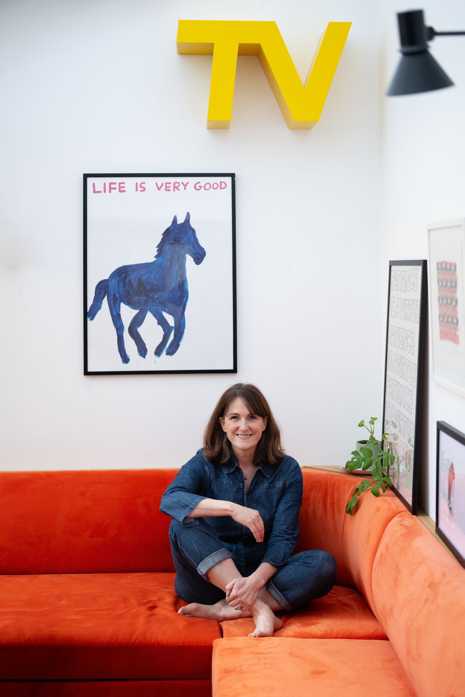 Woman in denim jumpsuit seated on orange sofa below yellow TV sign and horse artwork.