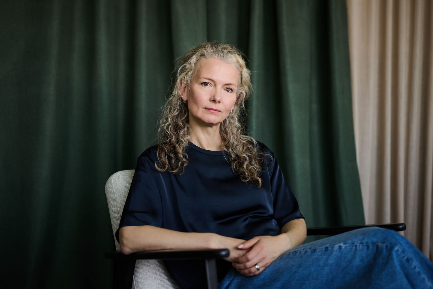 A woman with curly blonde hair sits in a chair wearing a navy top and jeans against a green curtain backdrop.