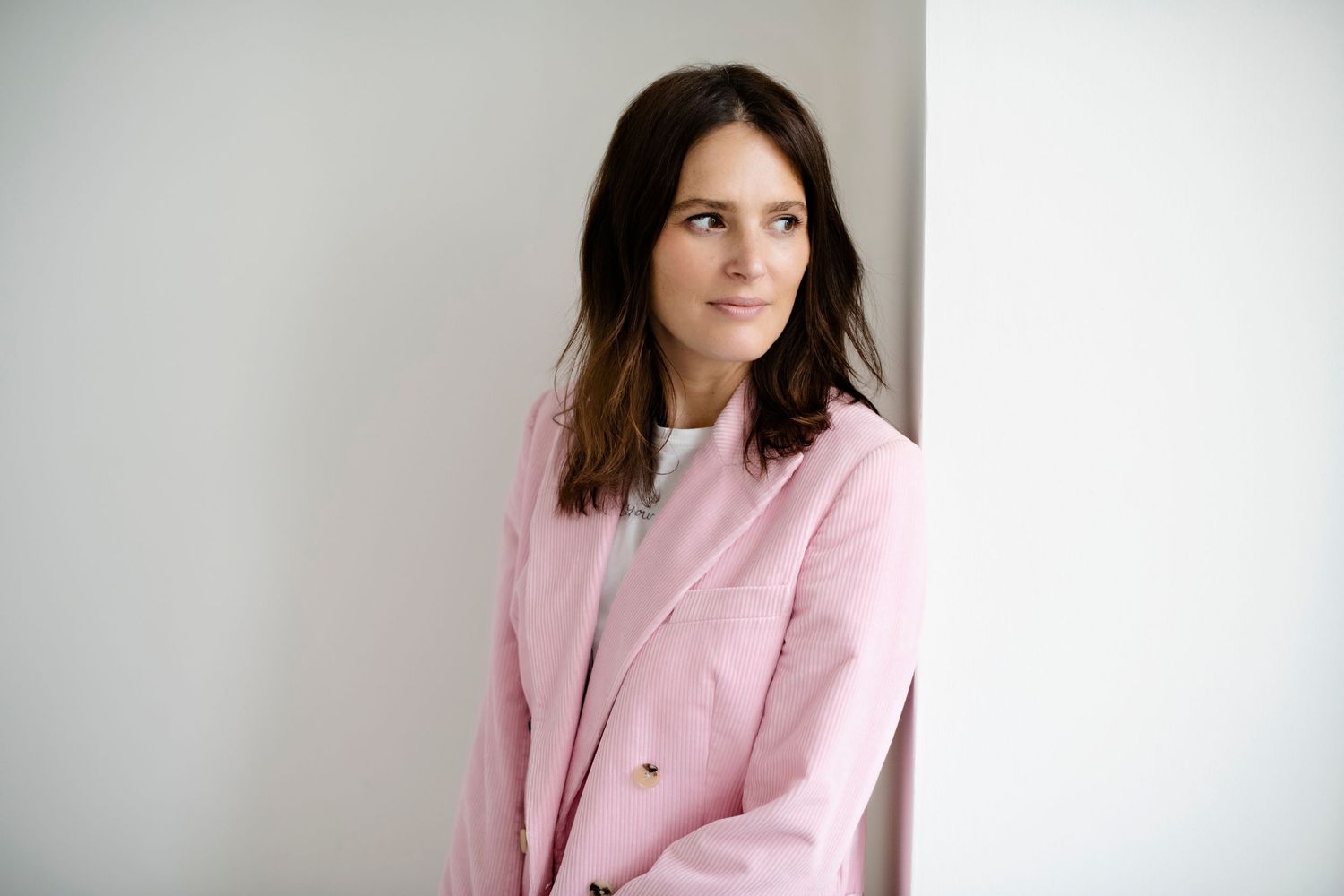A woman with dark hair wearing a pink blazer leans against a white wall, looking away thoughtfully.
