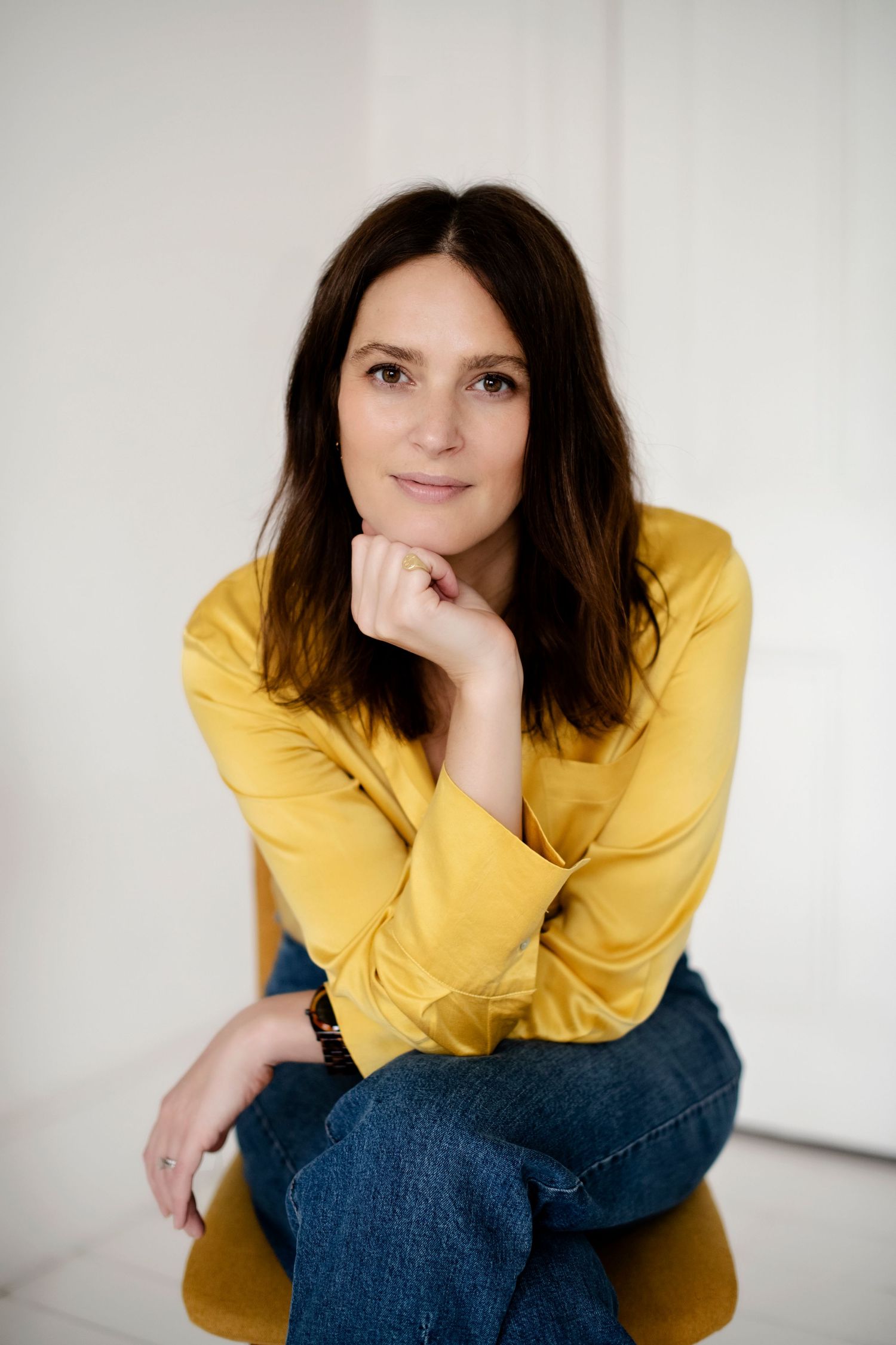 A woman in a yellow blouse and jeans sits relaxed on a chair, resting her chin on her hand against a white background.