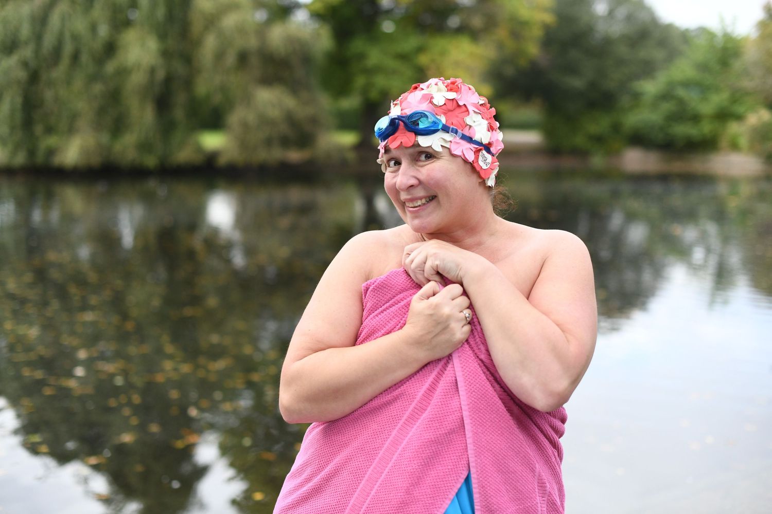 Woman in pink towel and swim cap smiles near outdoor lake after swimming.