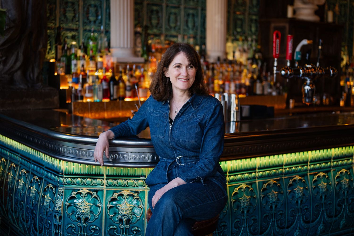 Woman in denim dress leaning on ornate tiled bar counter with bottles in background.