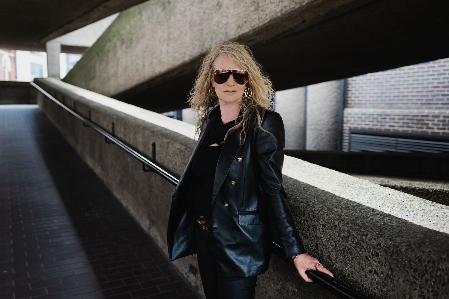 Blonde woman in black leather jacket and sunglasses posing in an urban underpass corridor.