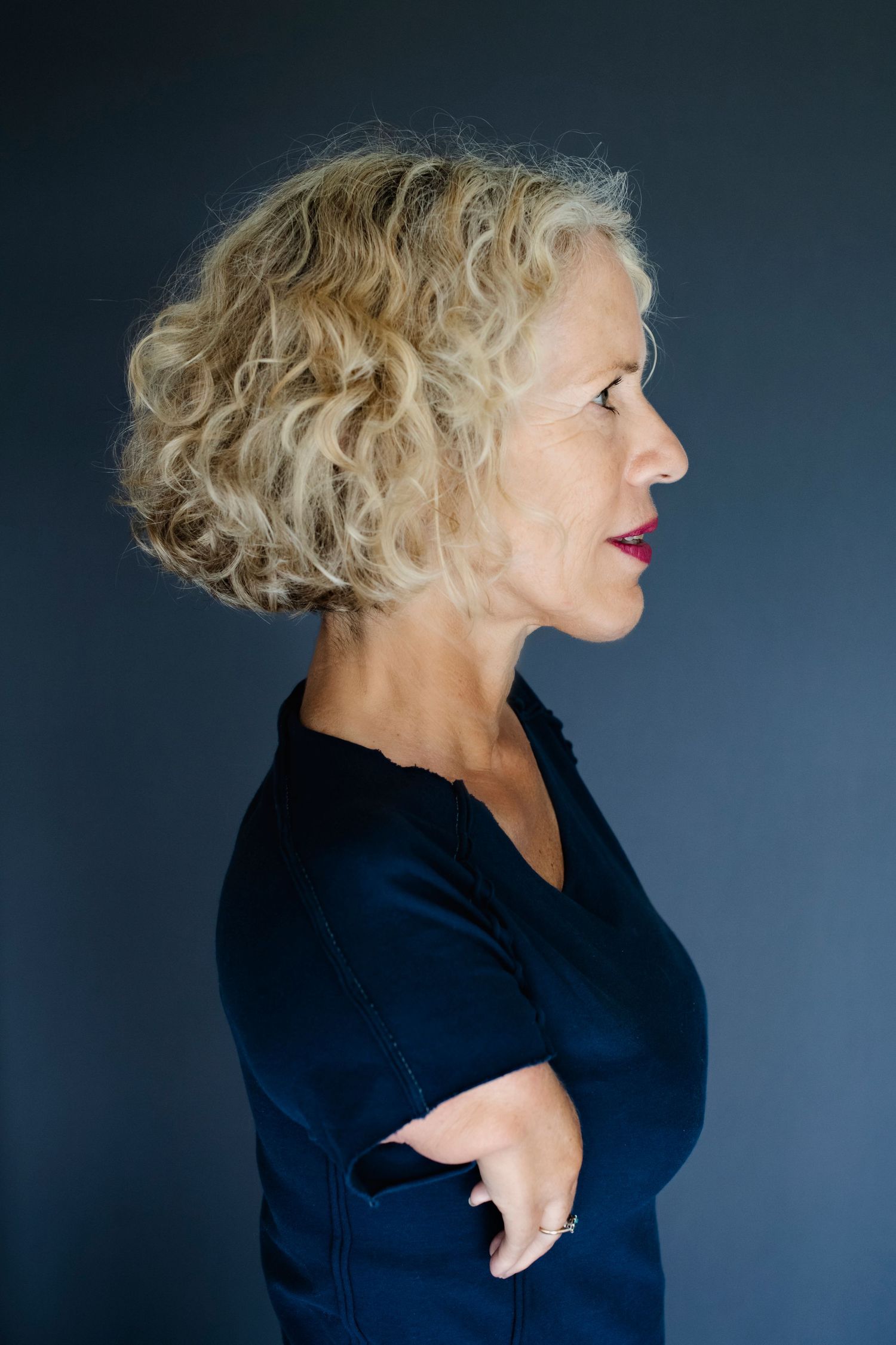 A side profile of a blonde curly-haired woman in navy with red lips and arms crossed on a dark gray background.