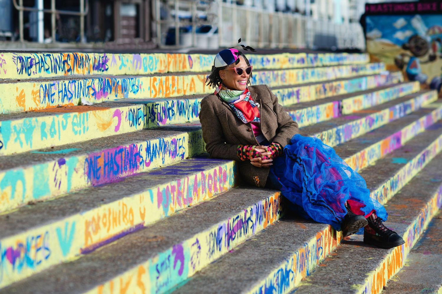 A colorfully dressed woman sits on graffiti-covered steps in a bright blue skirt and brown jacket.