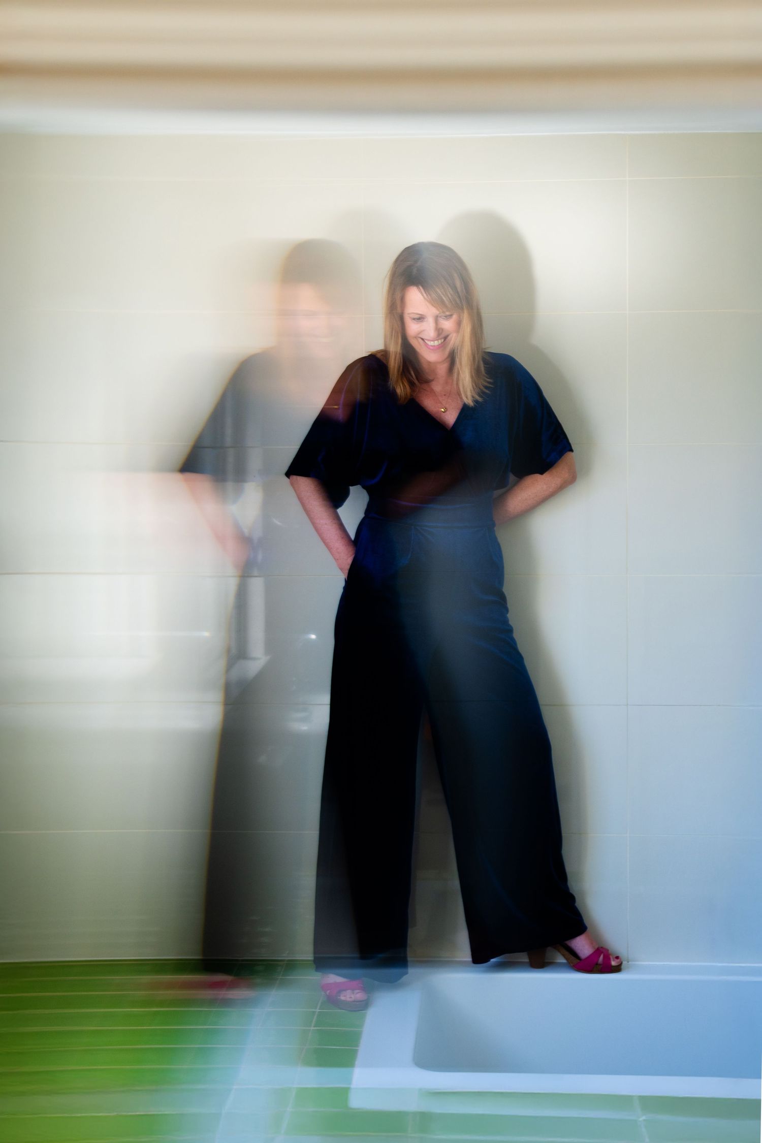 Motion blur artistic photo of woman in black jumpsuit standing in white bathroom with green tile.