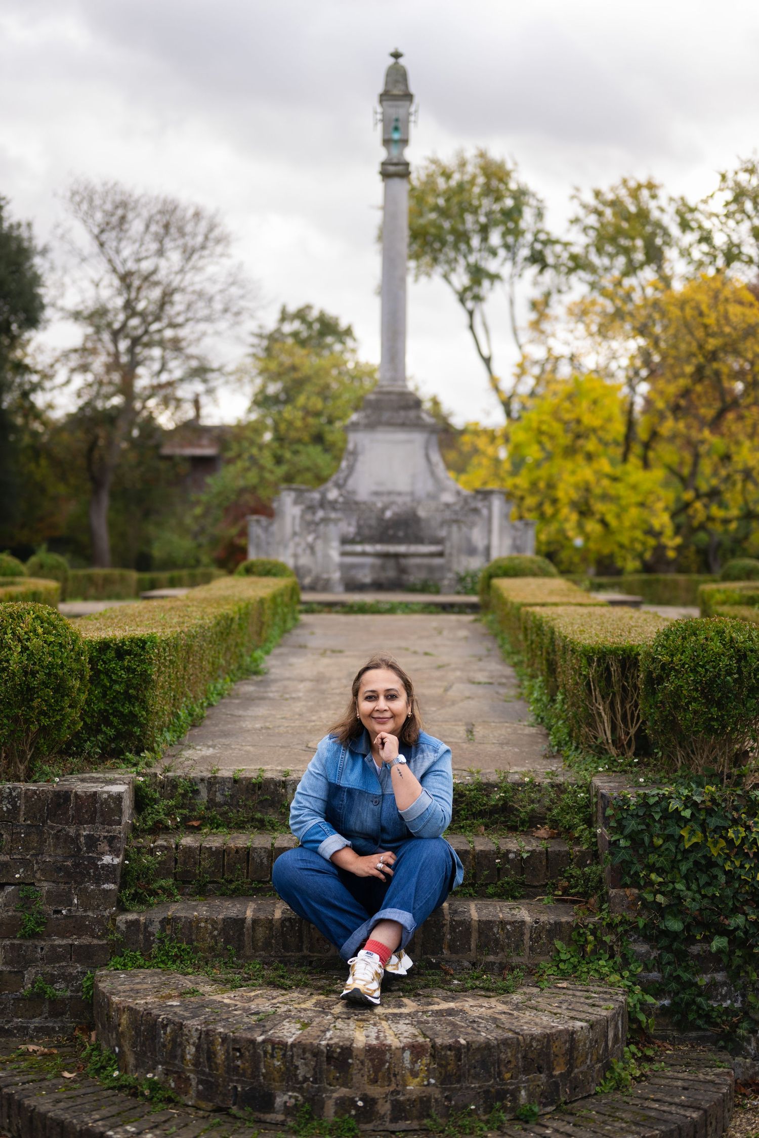 Woman in denim jumpsuit sitting on stone steps in a formal garden with a monument column backdrop.