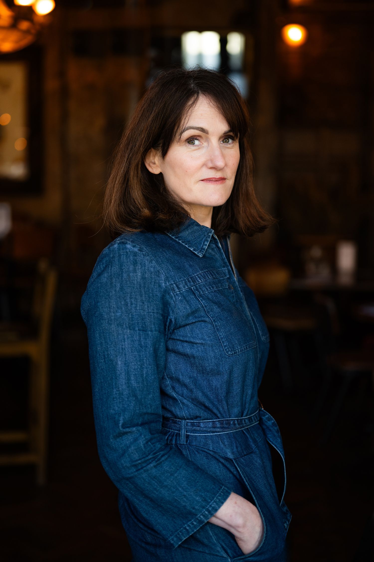 Woman in denim jumpsuit standing in dimly lit bar interior, looking over shoulder.