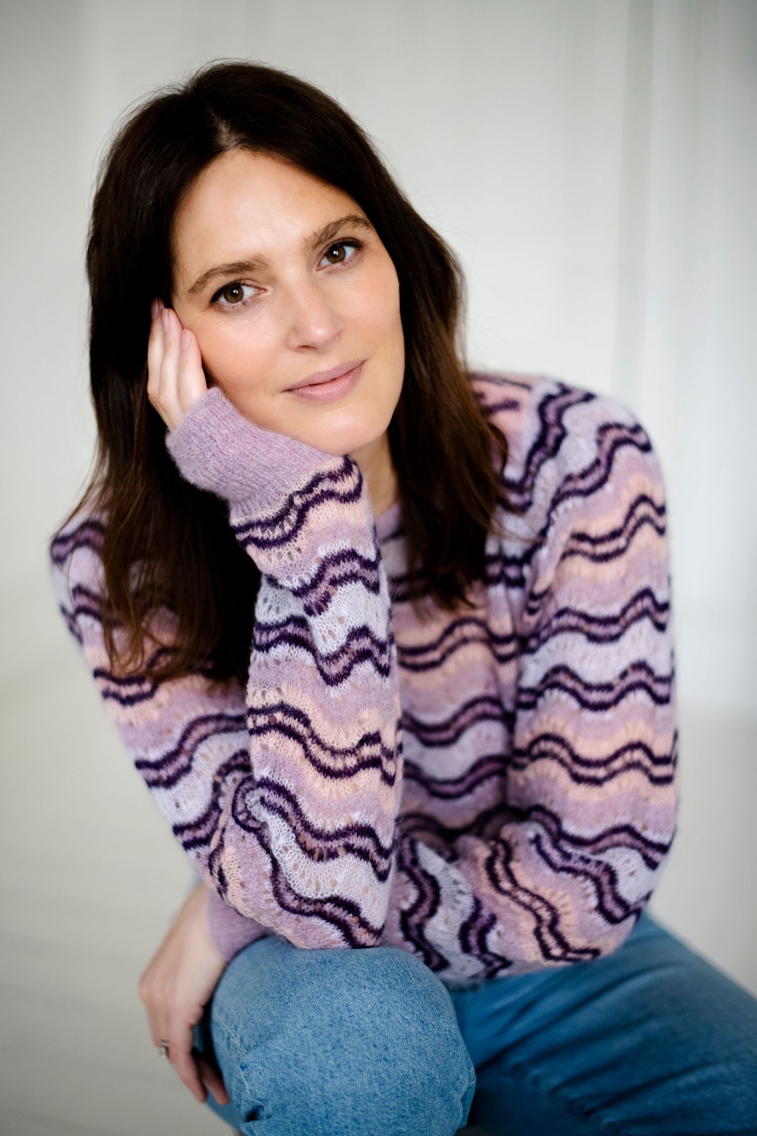 Brunette woman in a purple wave-patterned sweater and jeans posing with hand on cheek.