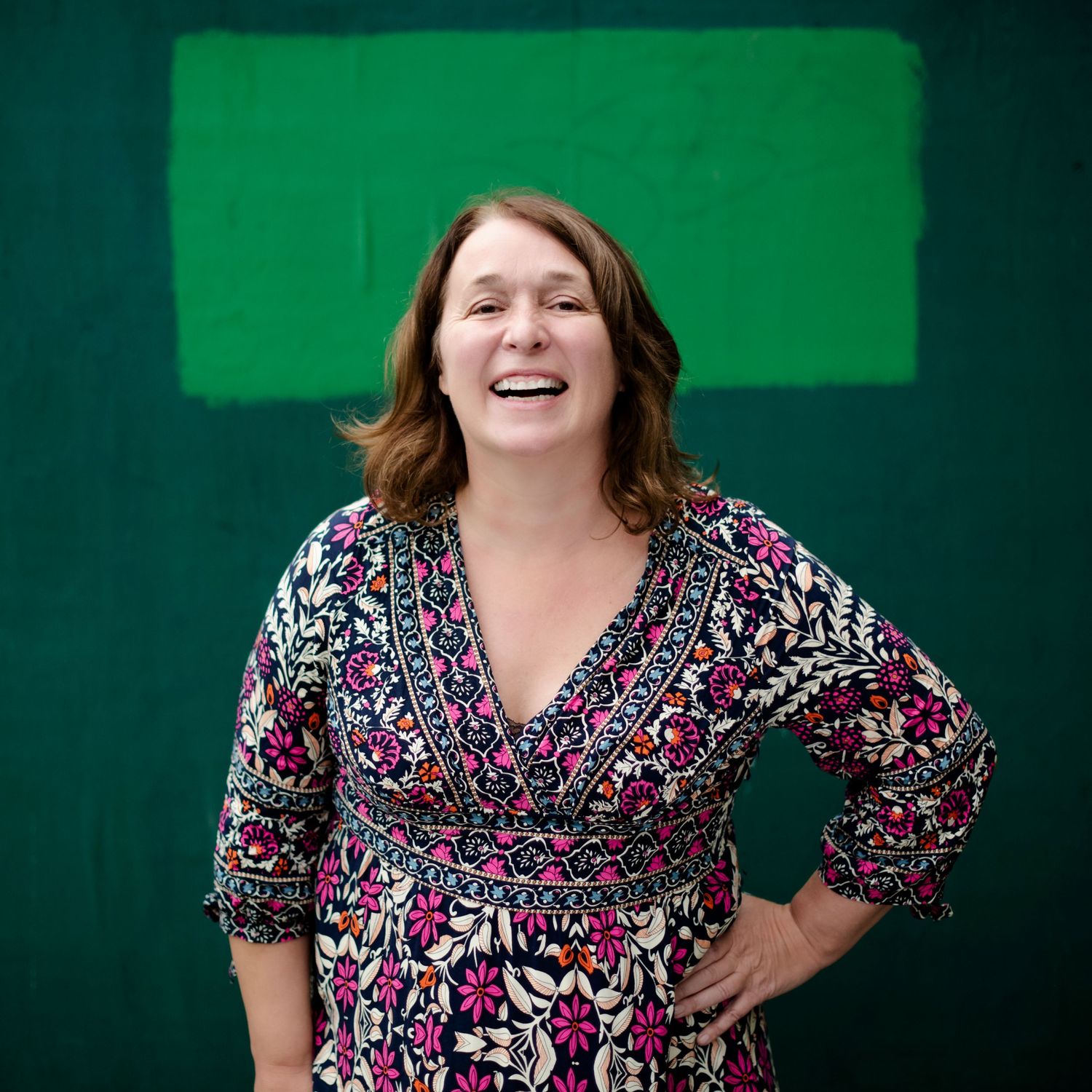 A smiling woman in a floral dress poses confidently in front of a green wall with a bright rectangle painting.