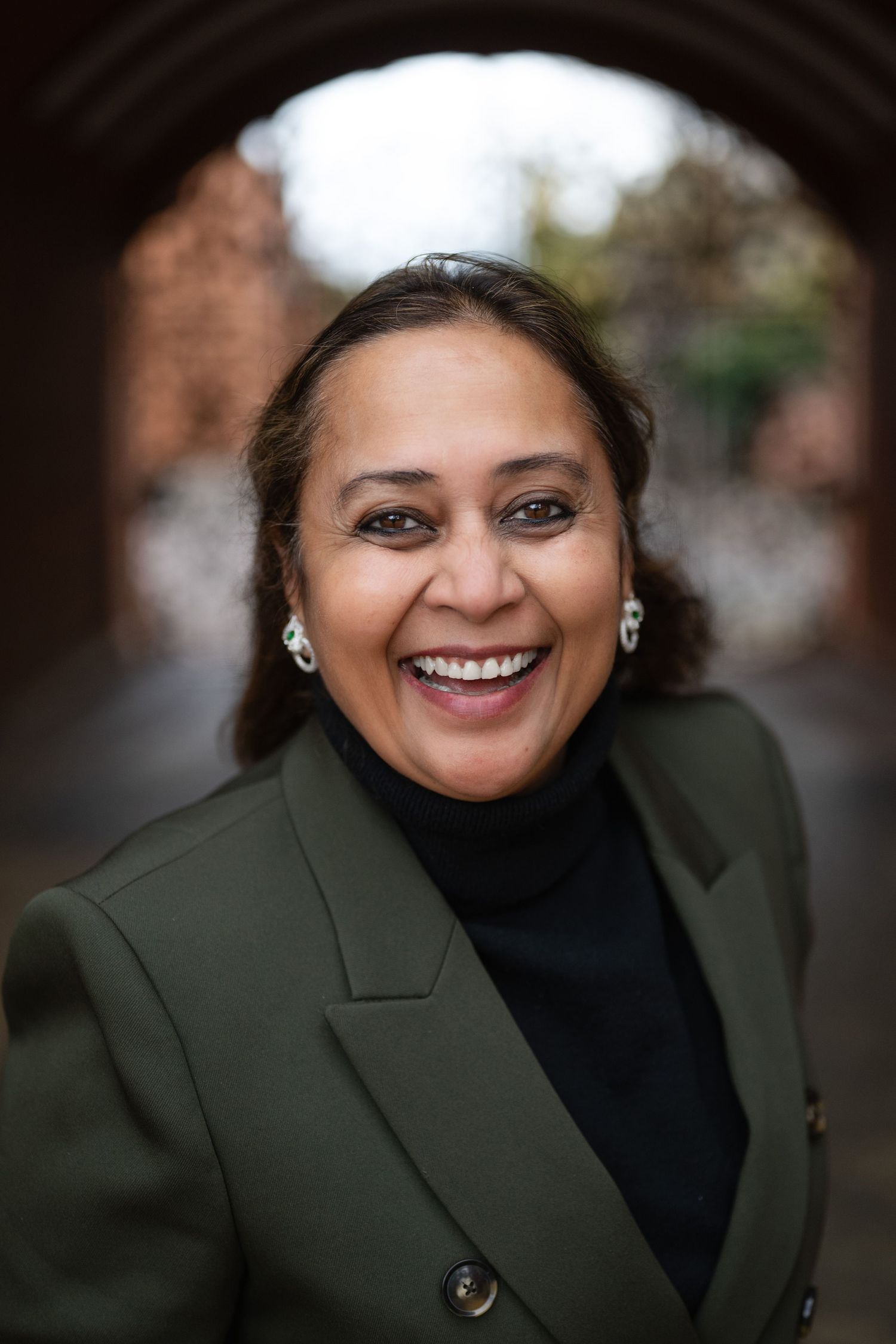 Smiling woman in green blazer and black turtleneck standing under a brick archway outdoors.