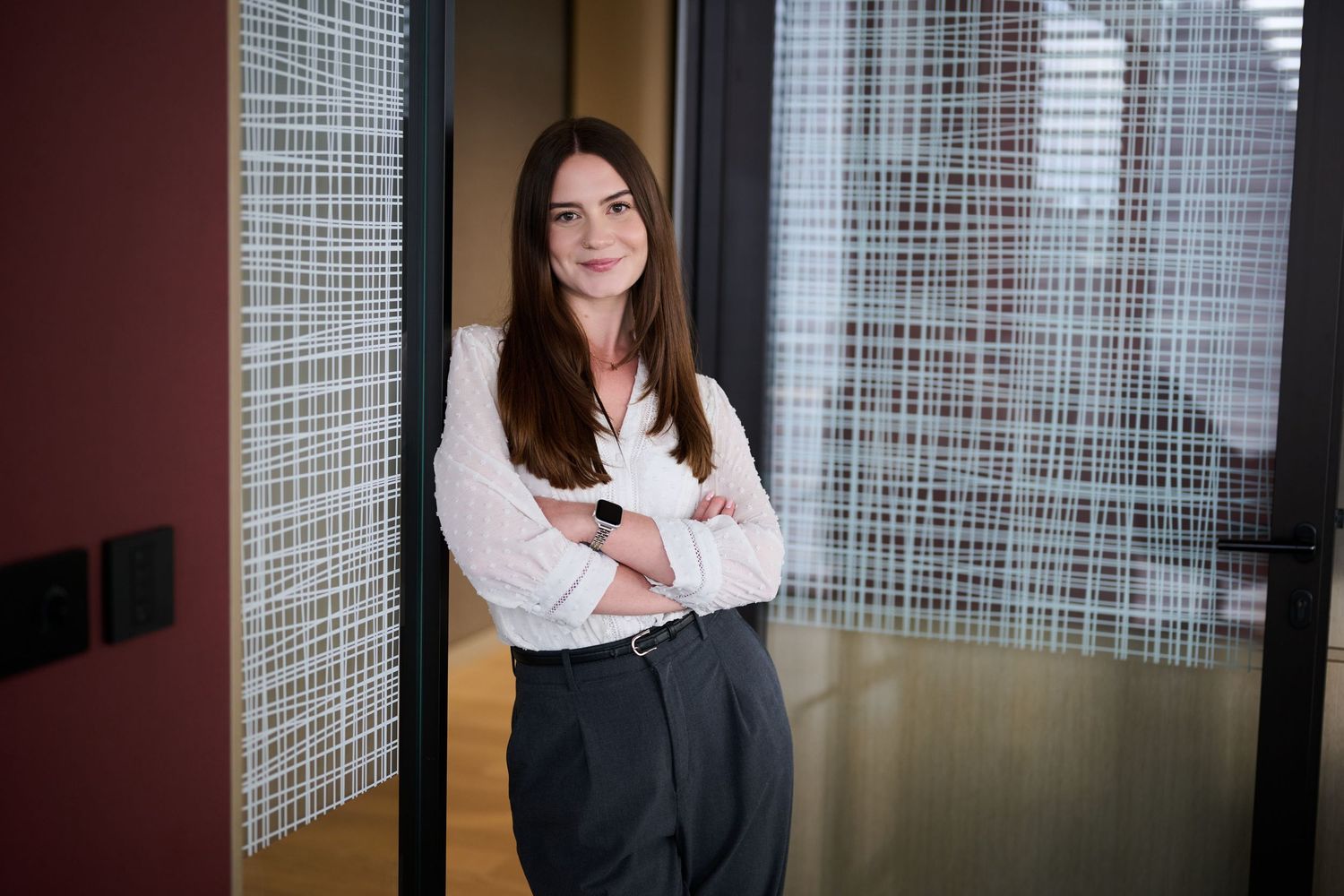 Young brunette woman in white blouse with arms crossed smiling in modern office hallway.