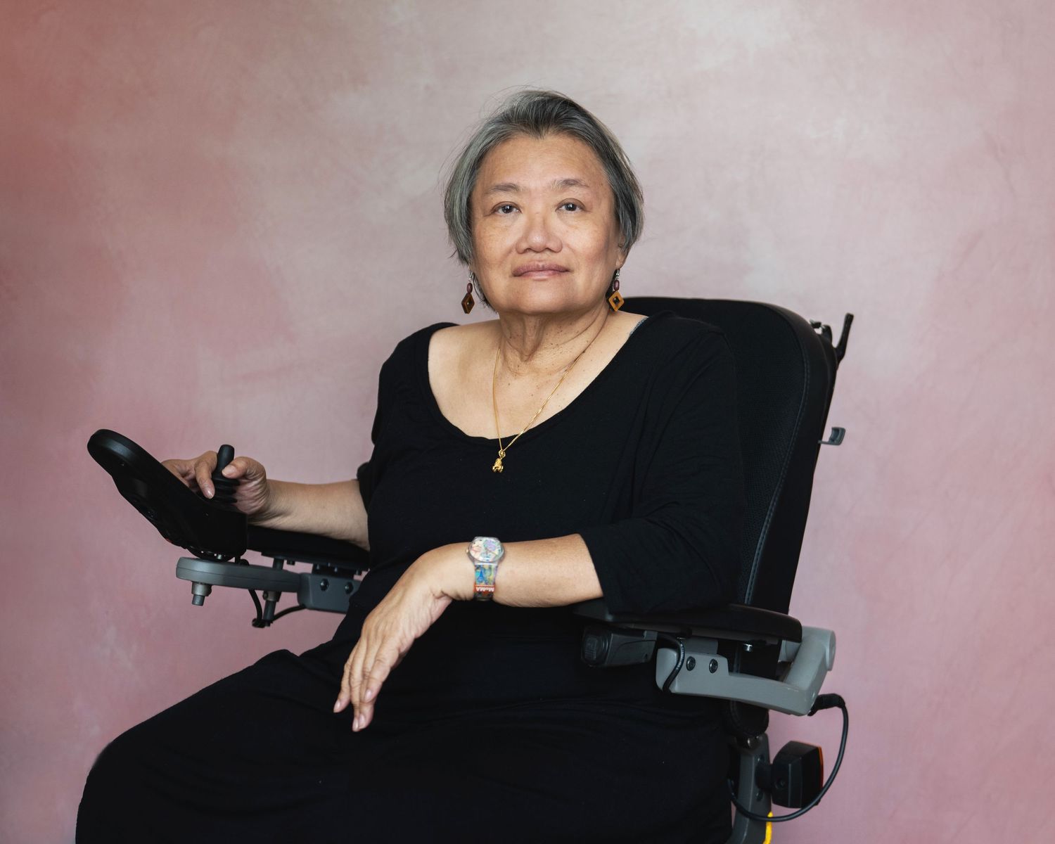 Woman in black outfit sits confidently in a power wheelchair against a soft pink background.
