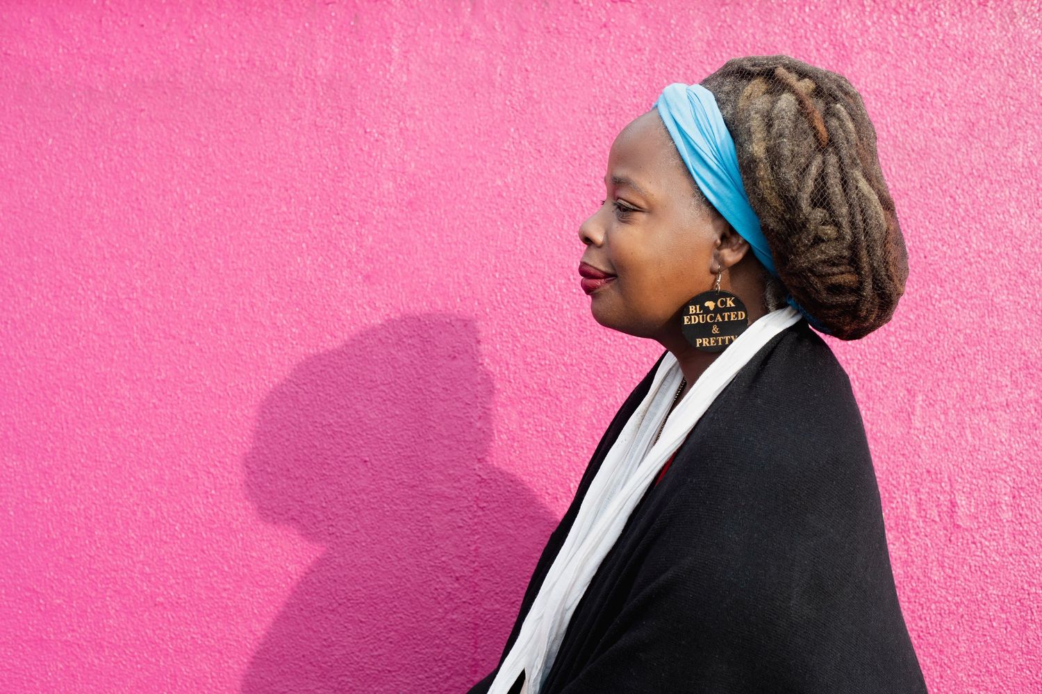 Woman with blue headband and locs stands in profile against vibrant pink wall.