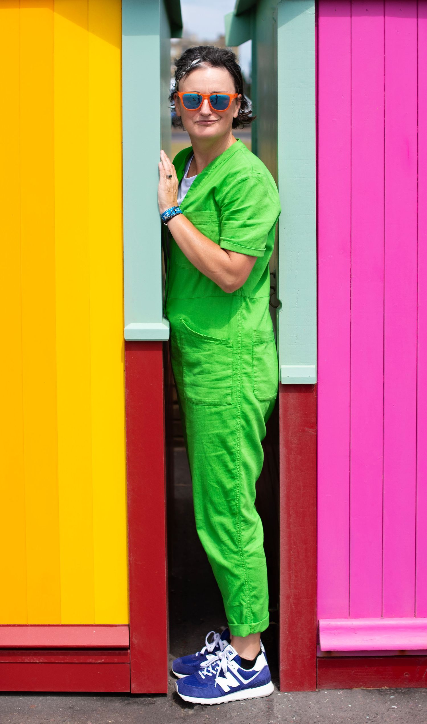 Woman in bright green jumpsuit and colorful sunglasses stands between vibrant yellow and pink painted walls.