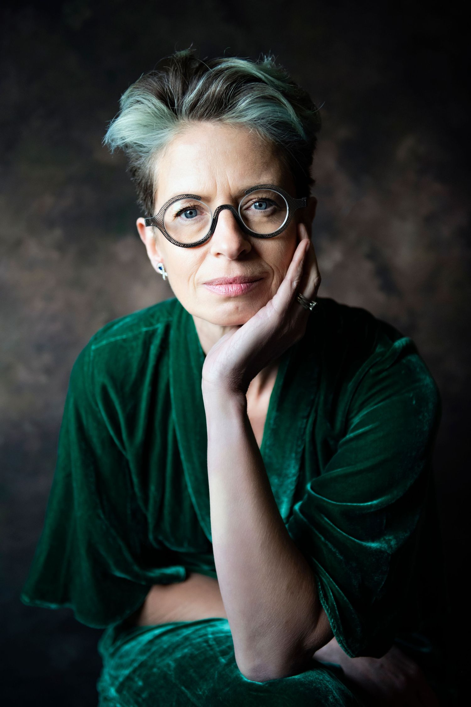 Woman with short gray-green hair and round glasses in a green velvet outfit against a dark background.