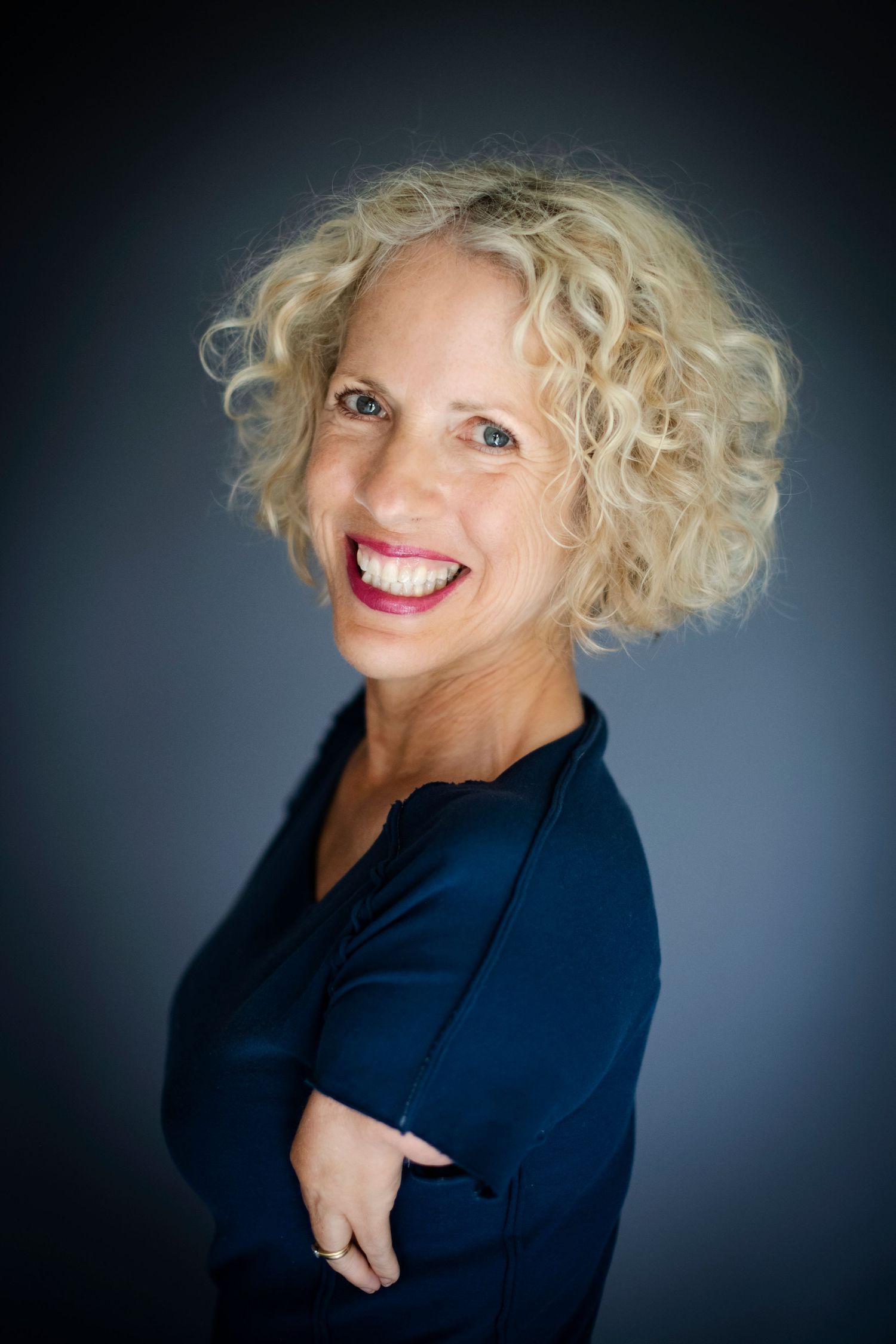 A curly blonde-haired woman in a navy top smiles brightly with arms crossed against a dark gray background.