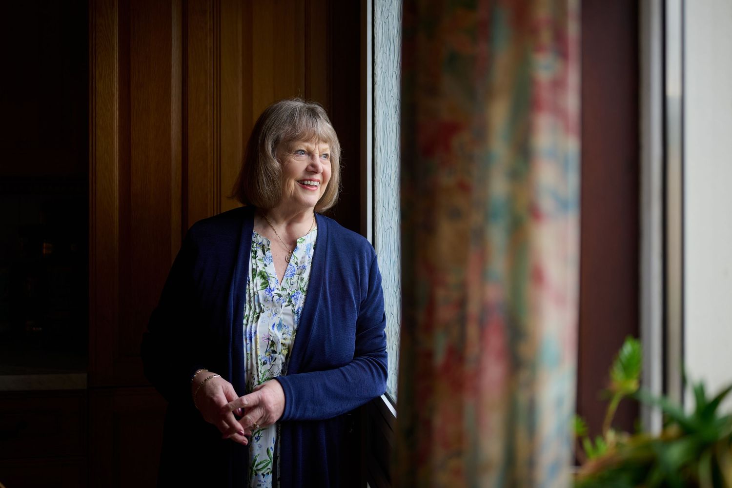Smiling older woman with short blonde hair stands by a window in soft natural light.
