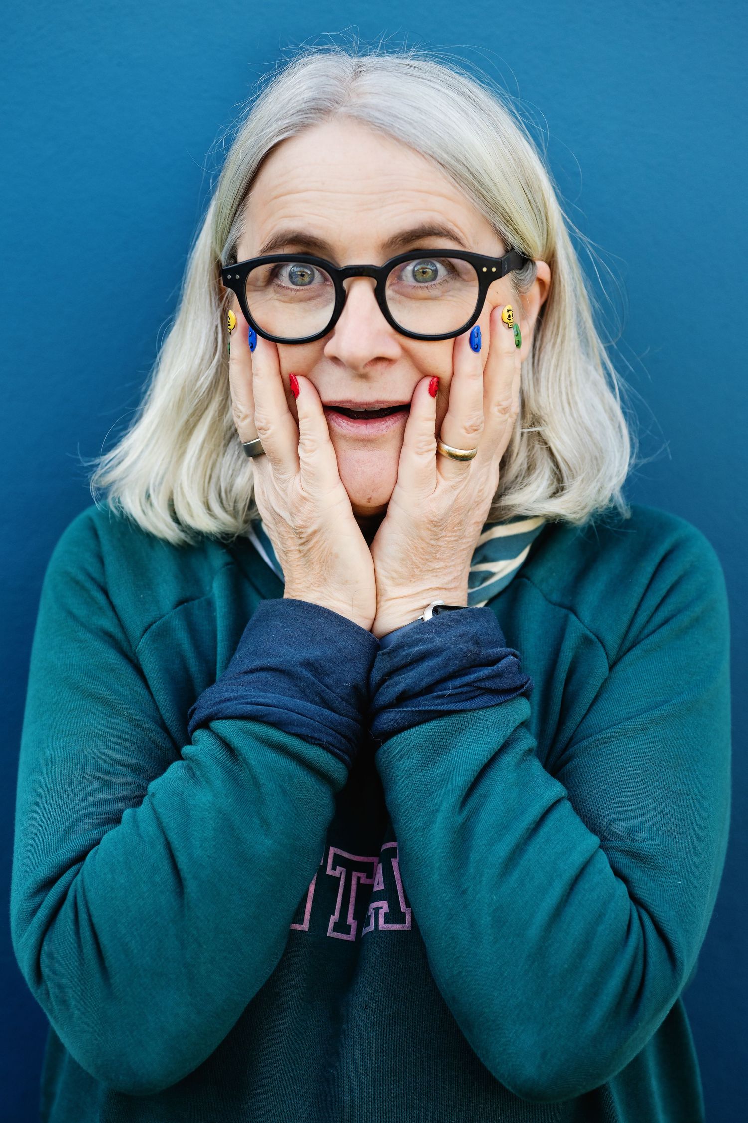 Surprised woman with gray bob and colorful nails holds face in hands against a blue background.